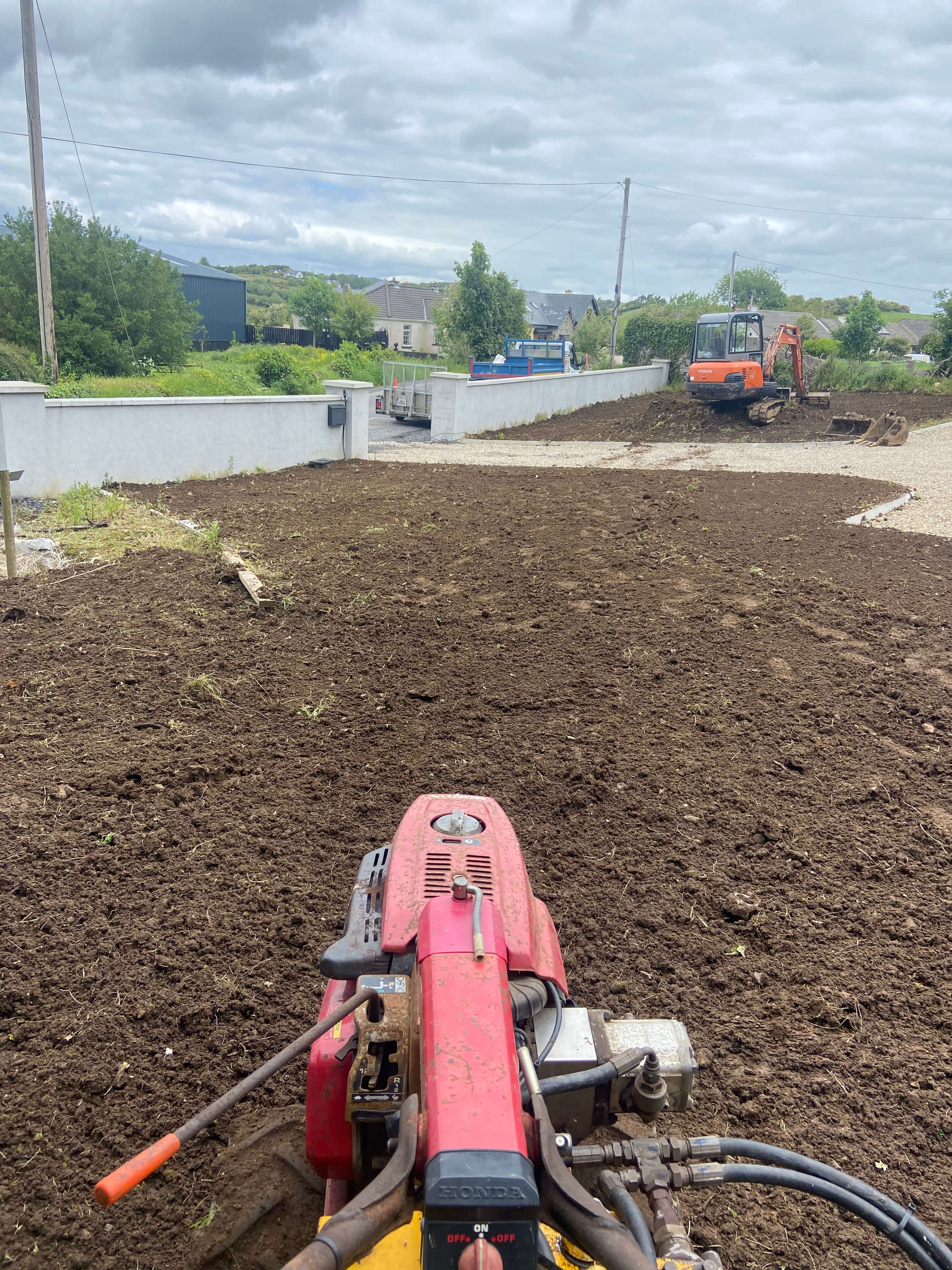View over freshly tilled soil with a red Honda rototiller in the foreground and a small orange excavator in the background near a white wall.