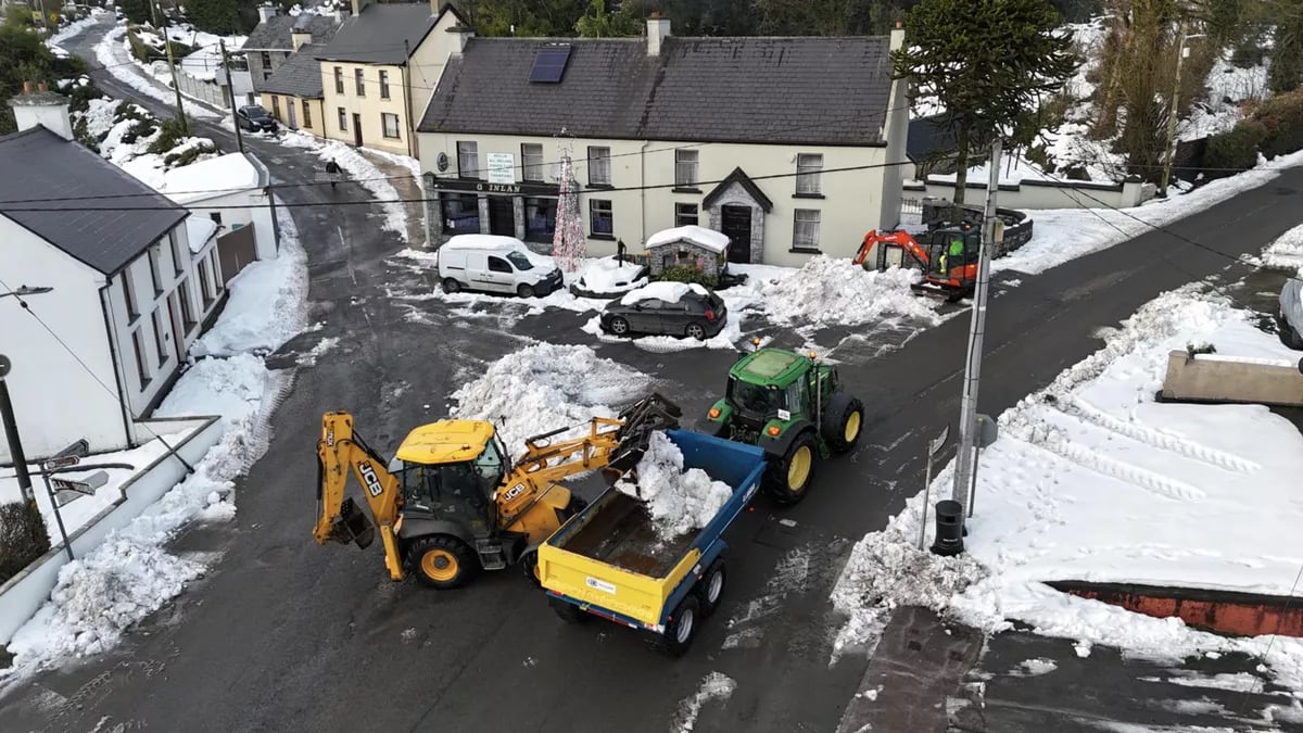 Snow removal work on a village street with a yellow JCB loader loading snow into a blue trailer attached to a green tractor, with snow-covered houses and roads.