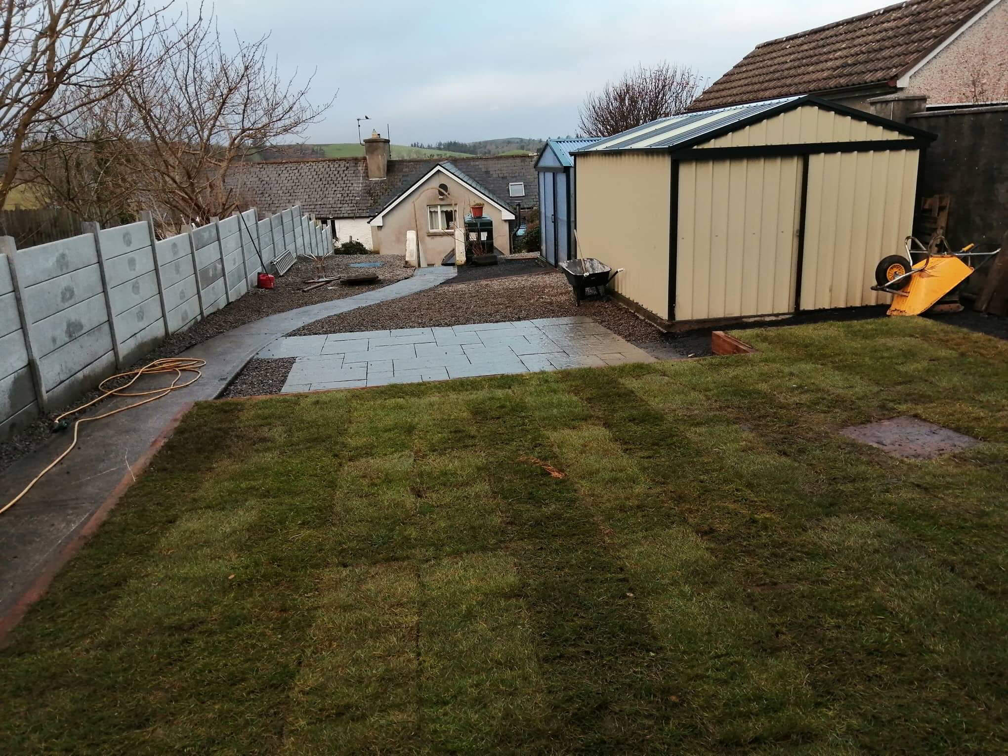 Backyard garden with freshly laid grass patches, concrete path, gravel area, beige garden shed, wheelbarrow, and a stone-paved section.