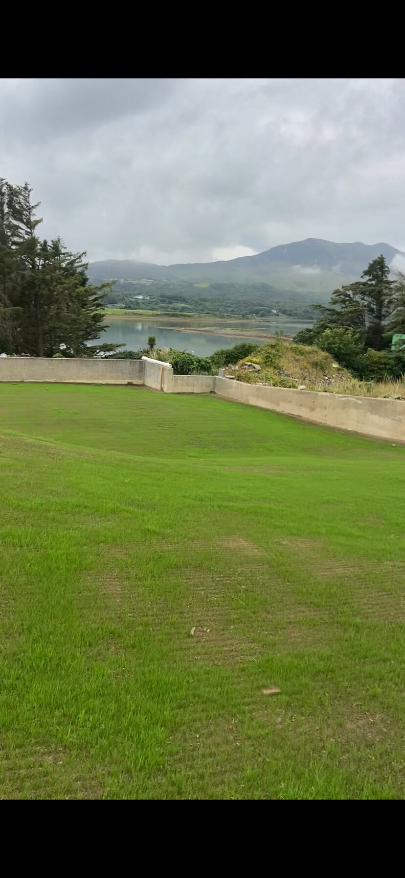 Green grassy lawn bordered by a low concrete wall with trees, water, and mist-covered mountains under a cloudy sky in the background.