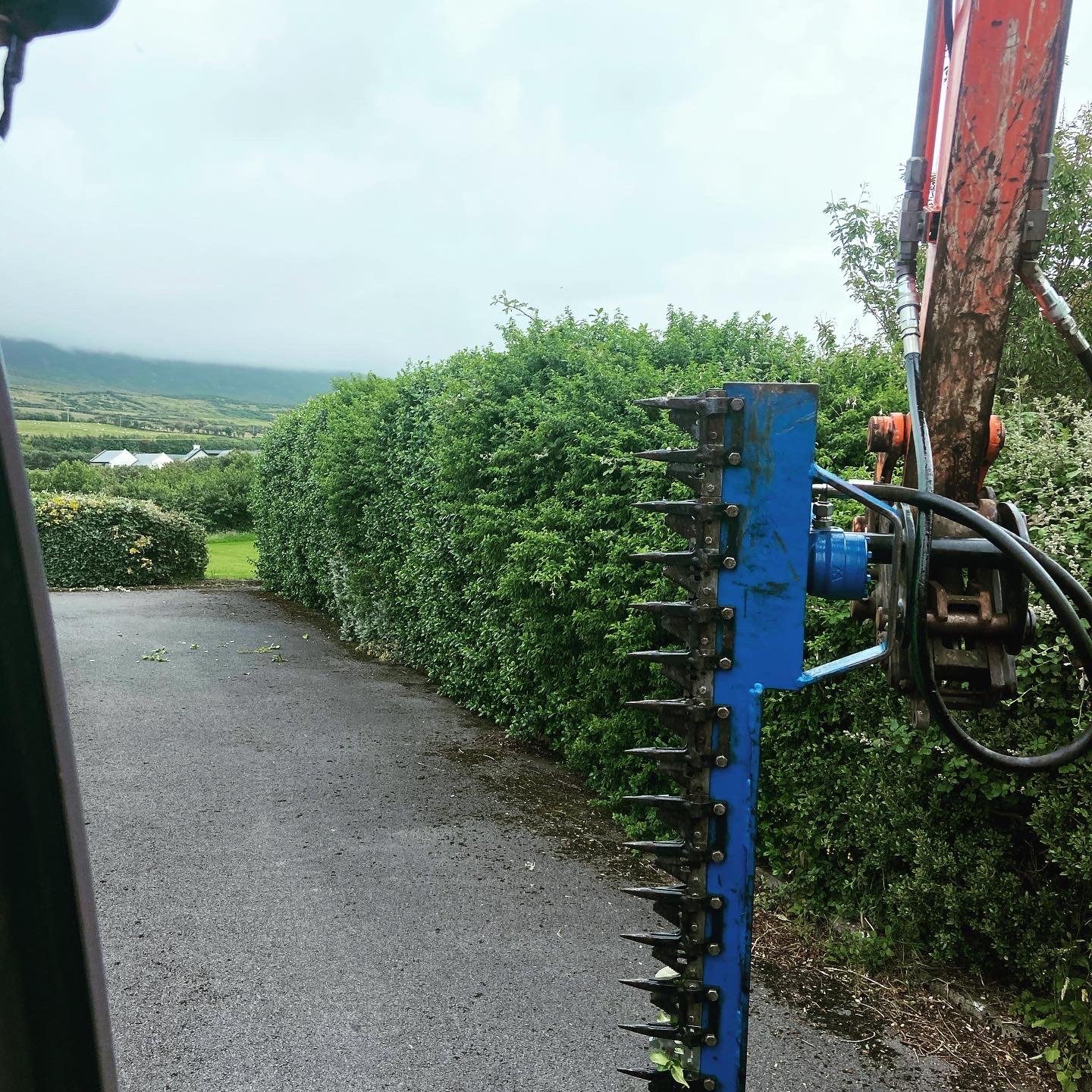 Close-up of a blue hedge trimming attachment on an orange excavator arm next to a neatly trimmed long green hedge by a paved driveway.