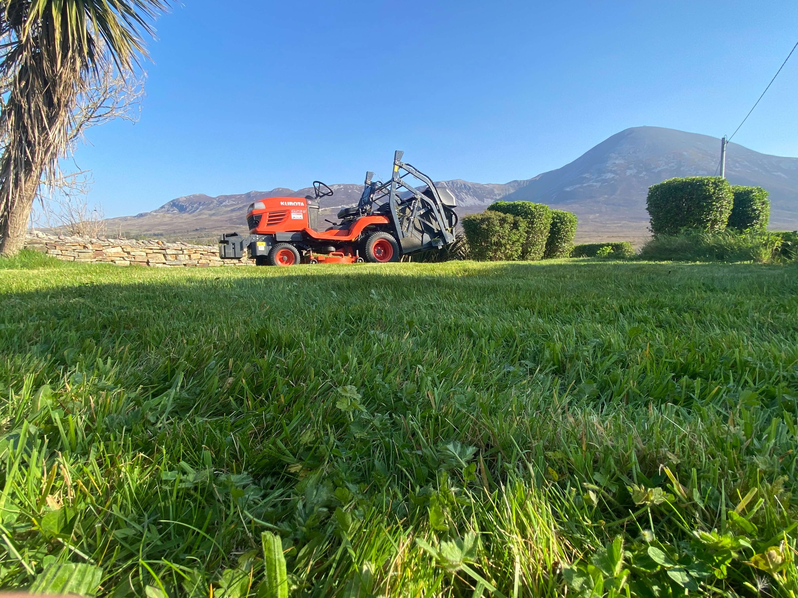 Red Kubota lawn tractor parked on green grass with trimmed shrubs and mountains in the background under a clear blue sky.