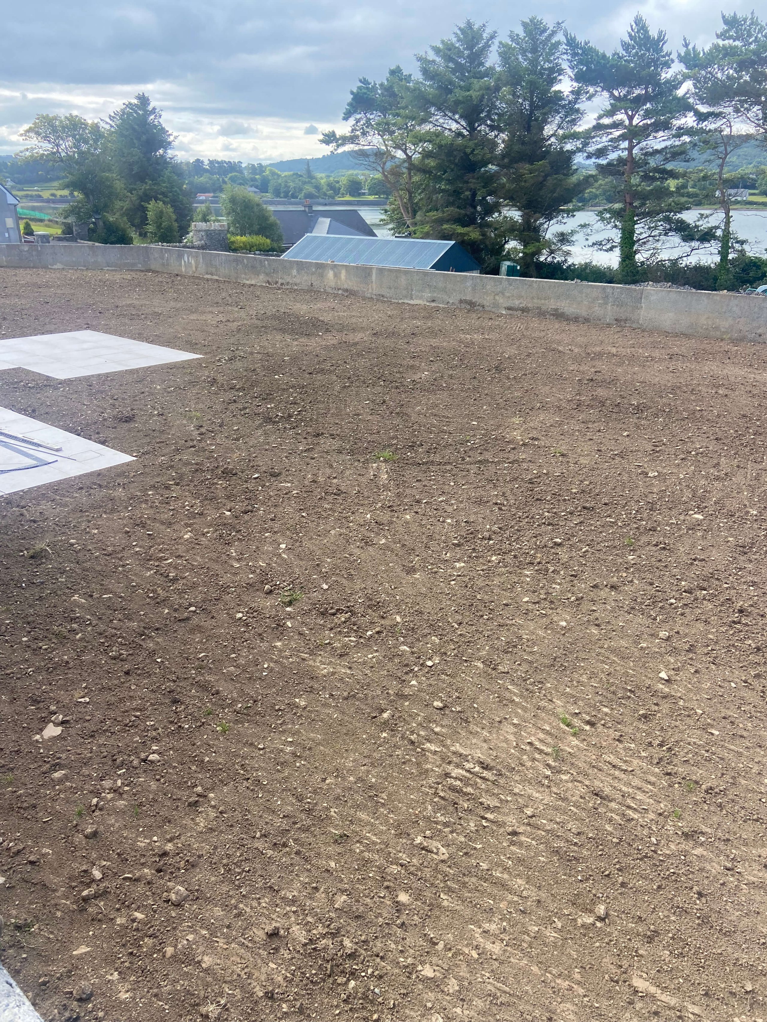 Cleared, leveled soil area bordered by a low concrete wall with trees, buildings, and a lake in the background under a cloudy sky.