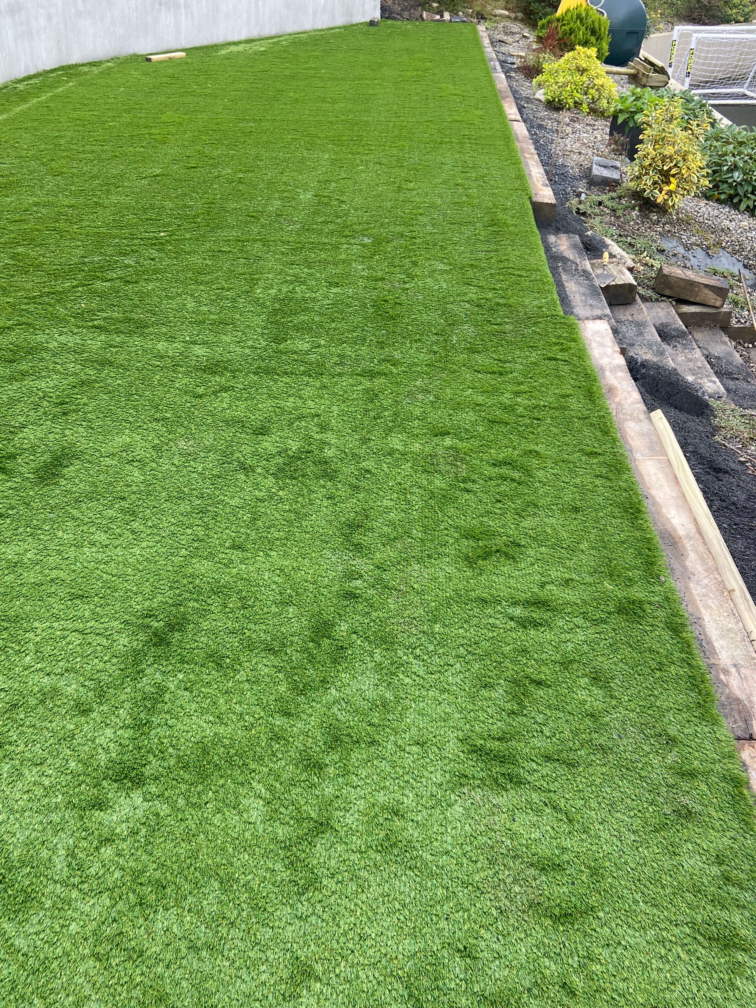 Newly installed artificial grass lawn bordered by wooden planks and adjacent to a gravel area with plants.