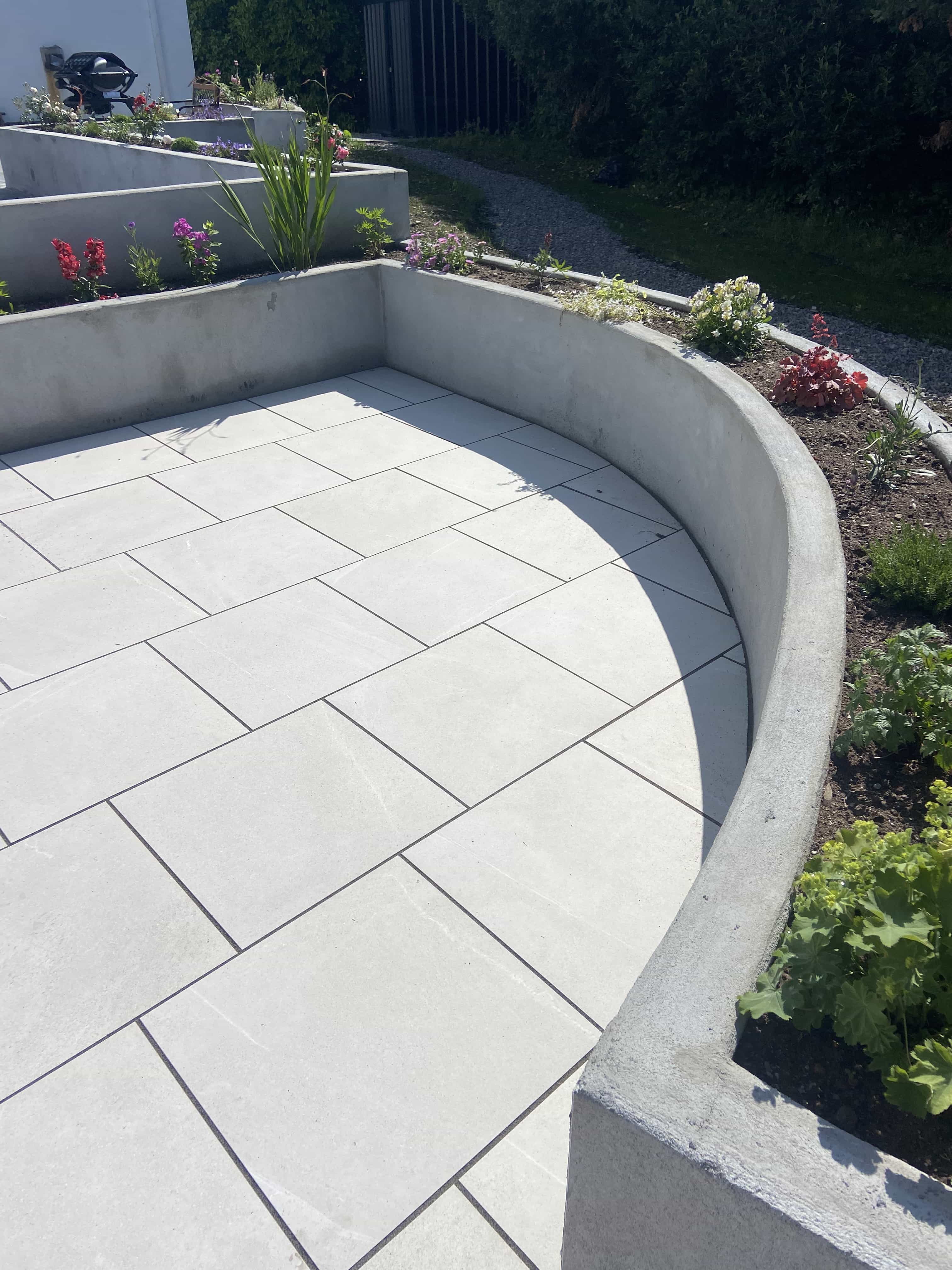 Curved outdoor patio with light gray tiles surrounded by low concrete planters with flowers and greenery.