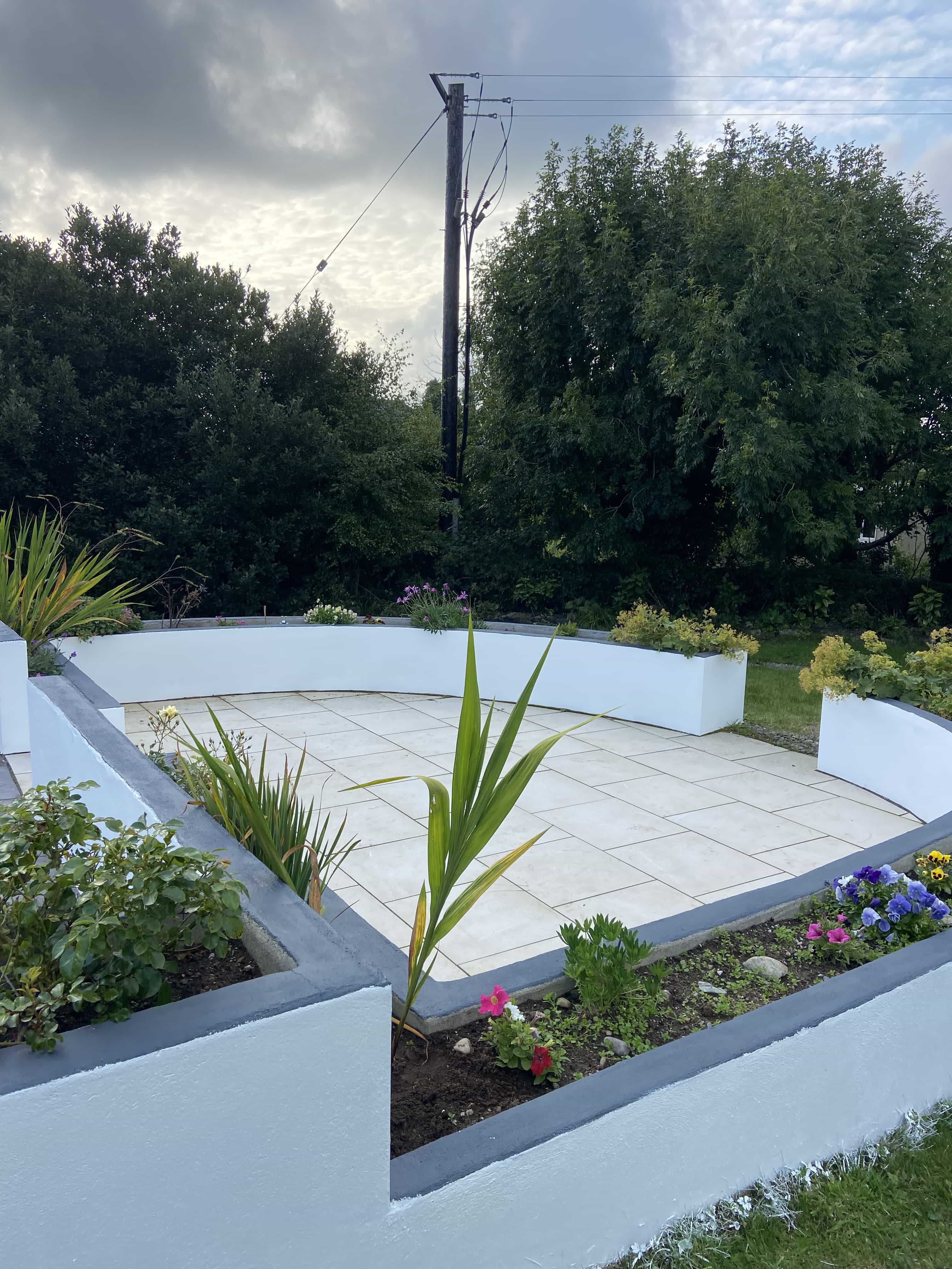 Curved white garden planters with gray trim surrounding a tiled patio, filled with various green plants and flowers, with tall trees and a utility pole in the background.
