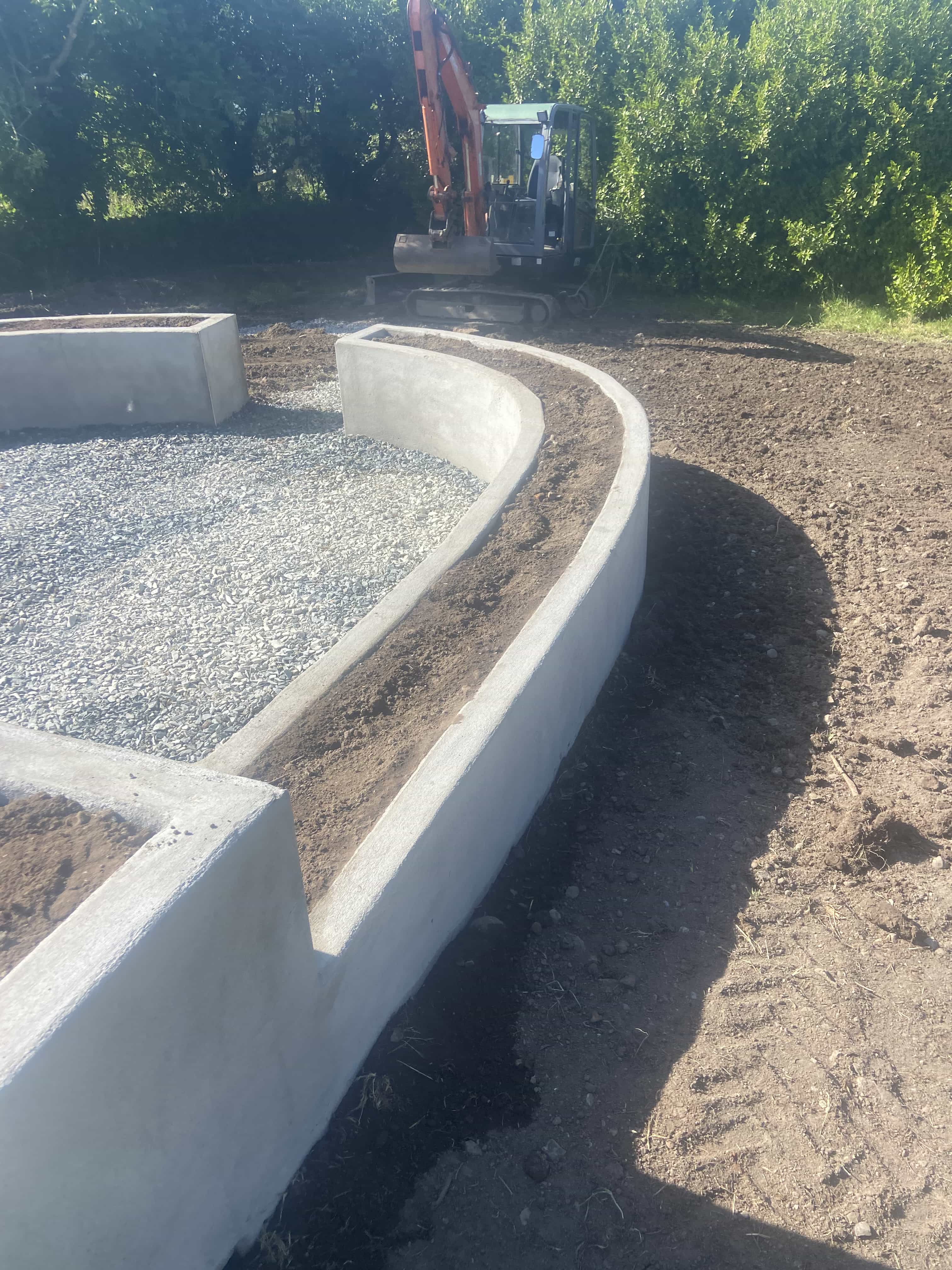 Curved concrete garden bed walls filled with gravel and soil, with a small excavator in the background.