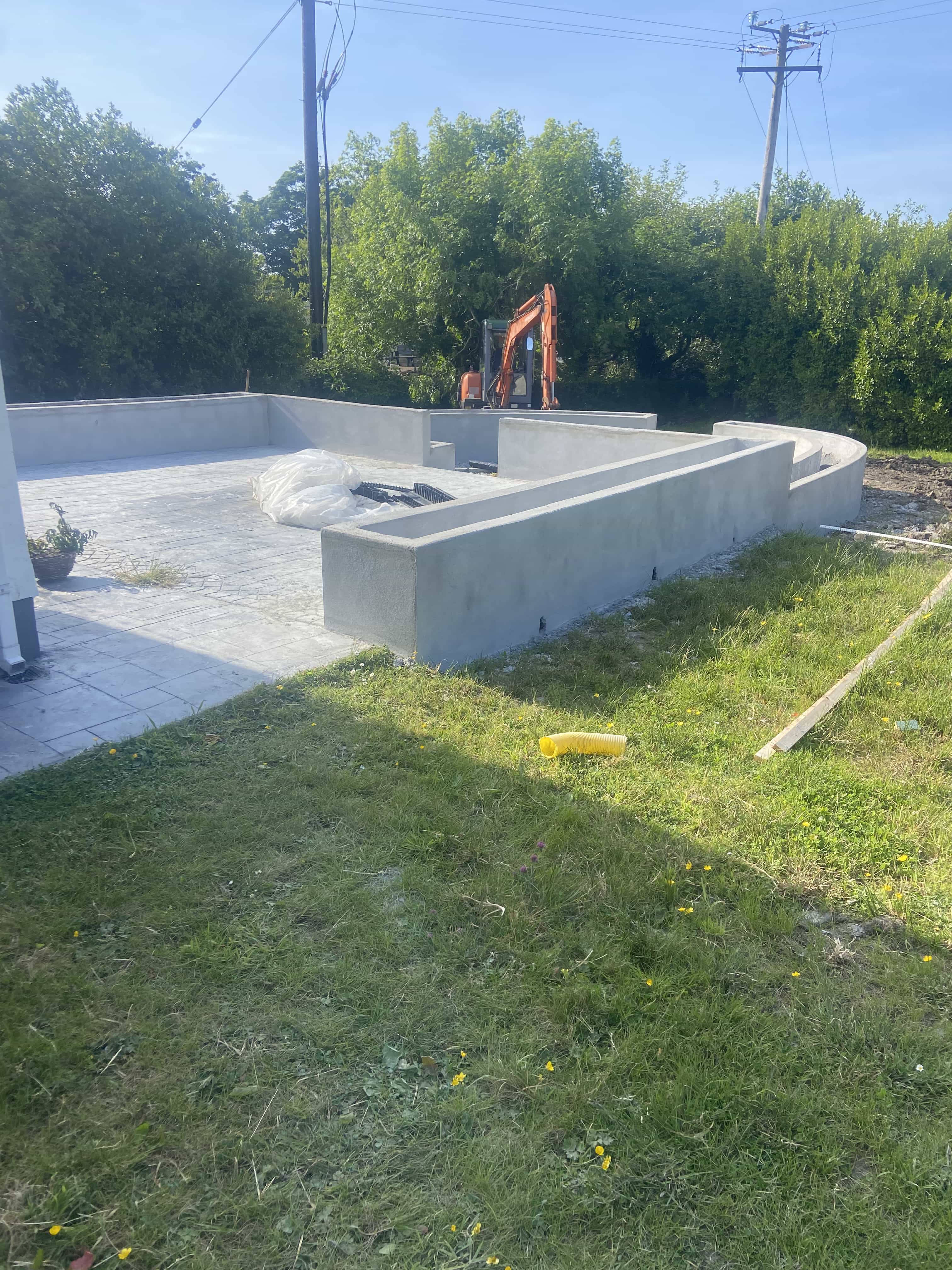 Construction site of a concrete patio with low walls, next to green grass and trees, with an orange excavator in the background.