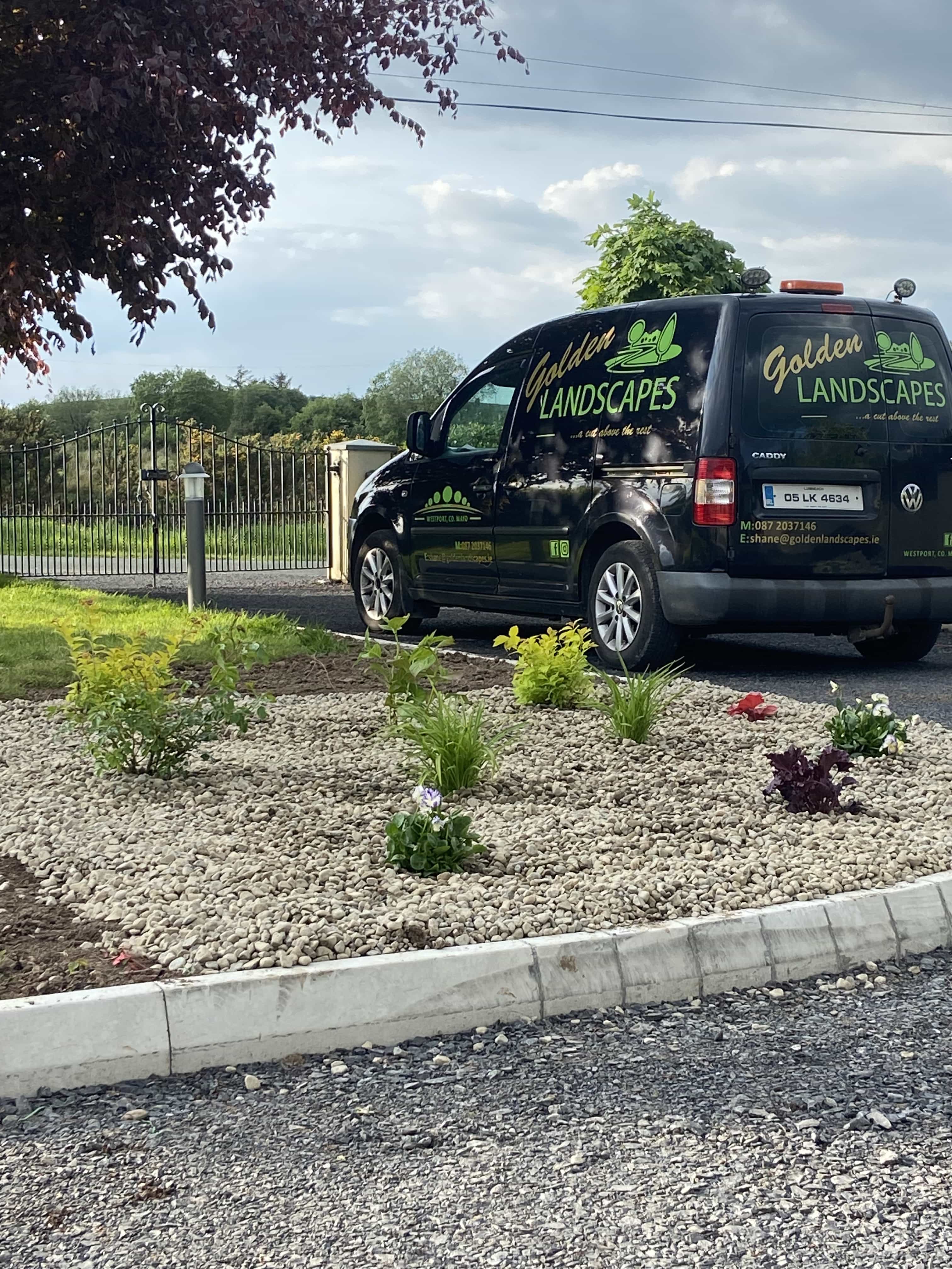 Landscaping van parked behind a newly landscaped area with white stones, small plants, and greenery under a partly cloudy sky.
