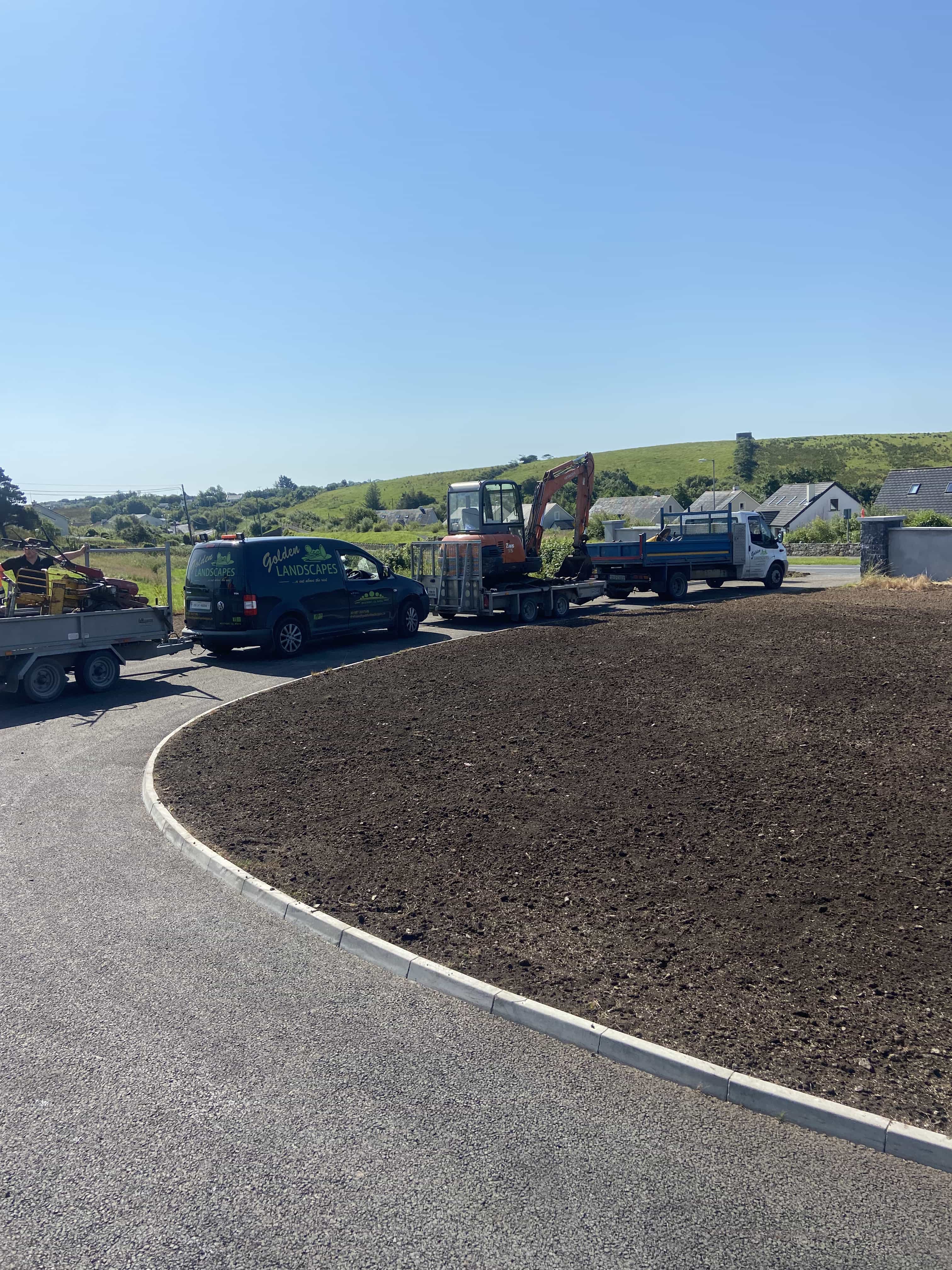 Landscaping vehicles and equipment parked beside recently tilled soil on a curved roadside under a clear blue sky.