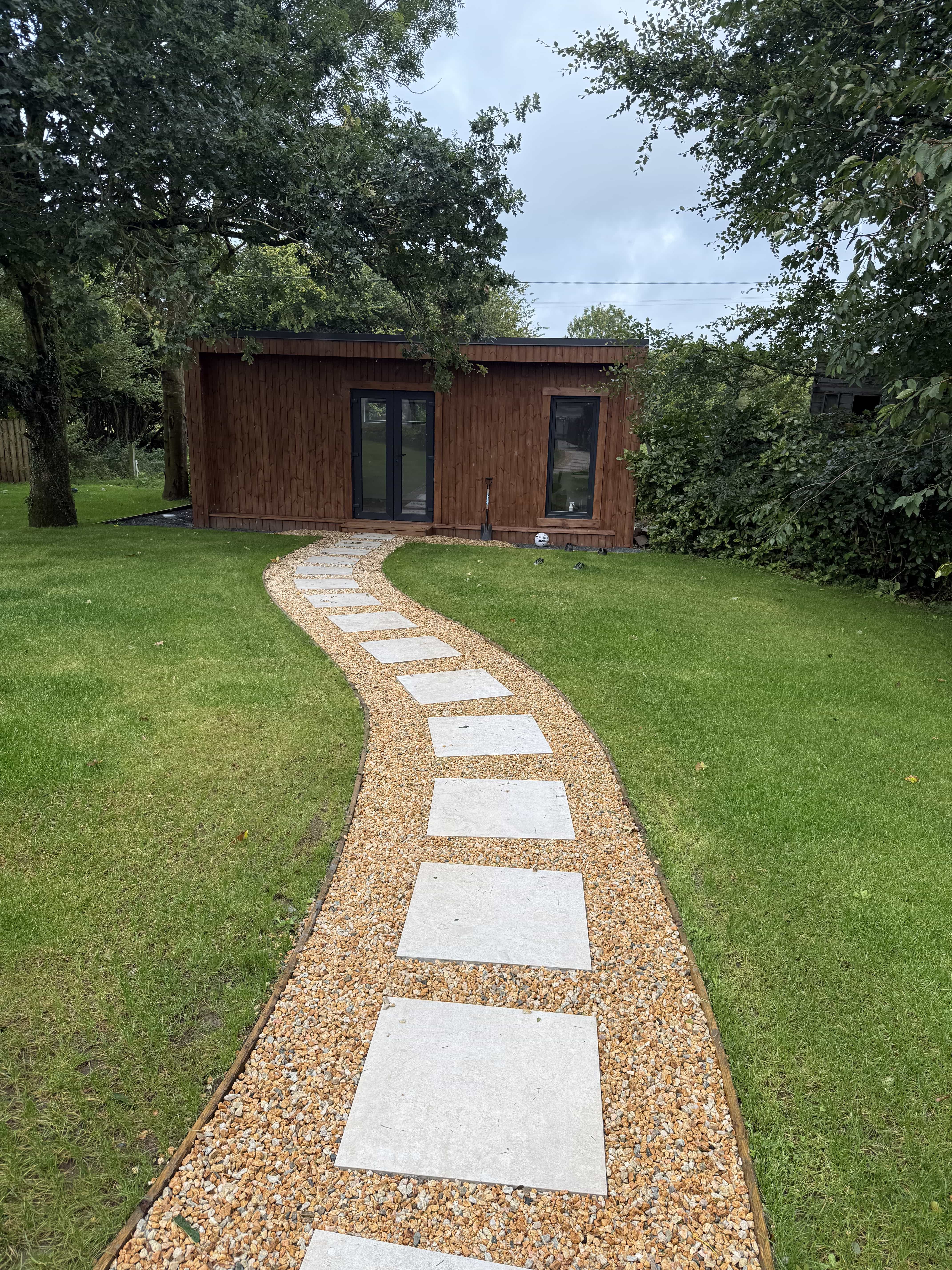 Stone stepping path curving through green lawn leading to a brown wooden garden shed with glass doors and windows.