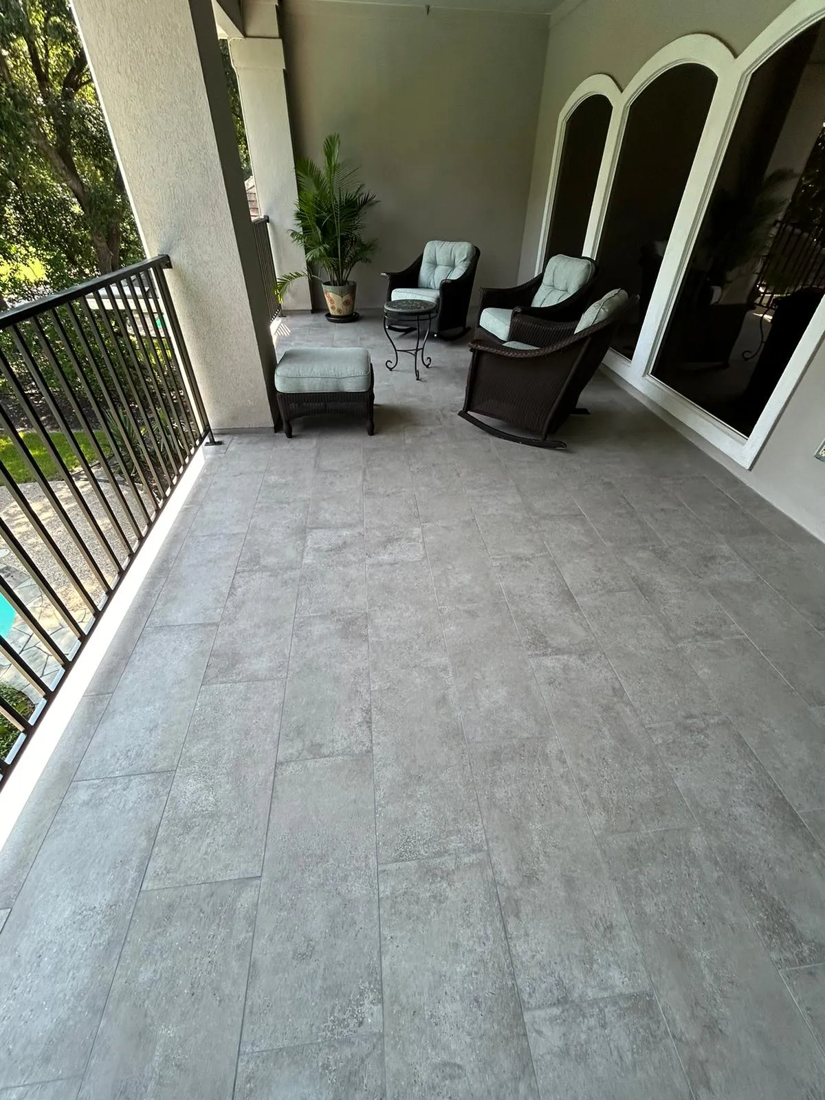 Covered patio with gray tiled flooring, wicker chairs with light cushions, a small glass table, and a potted plant near a railing overlooking greenery.