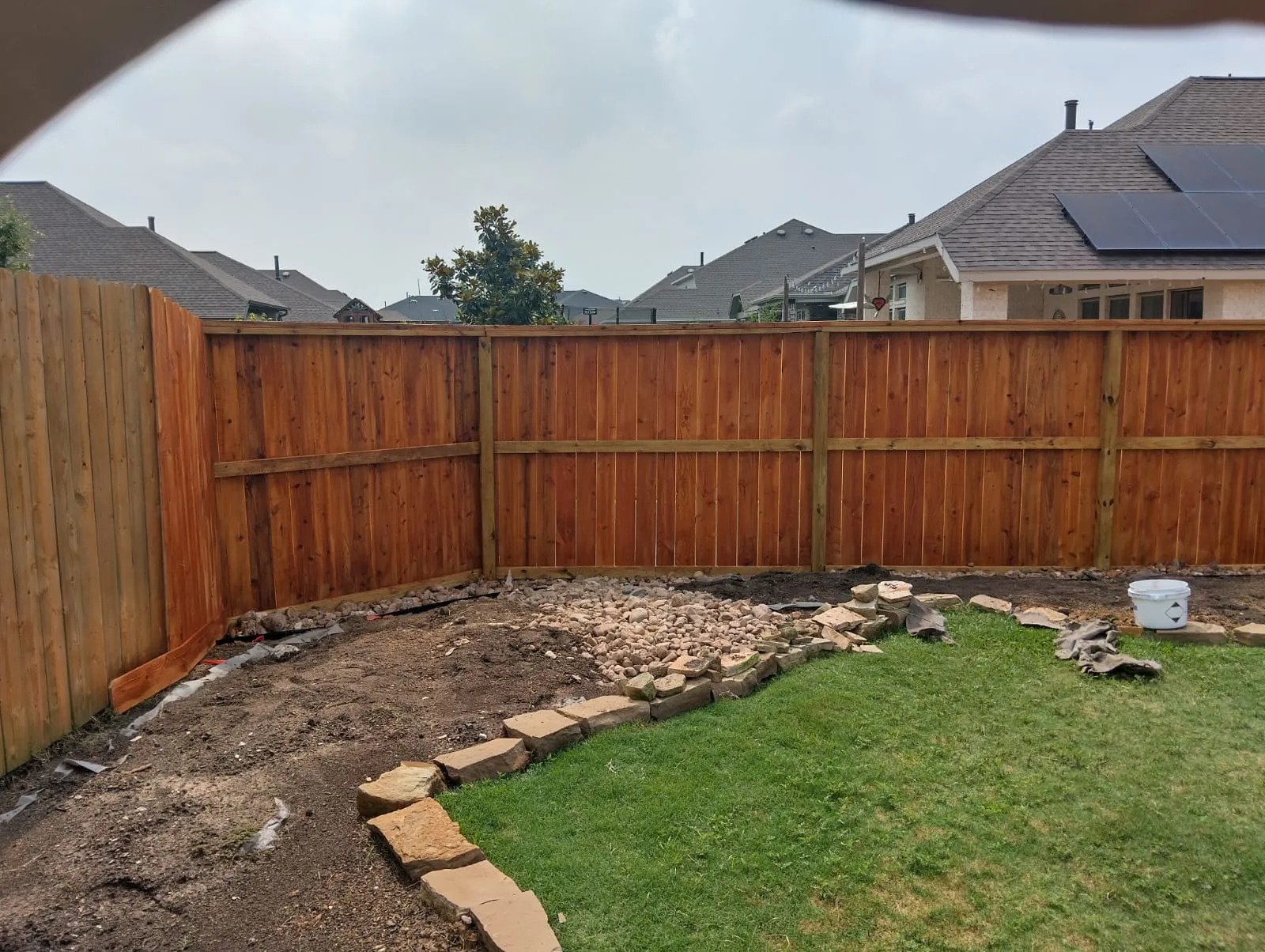 Backyard with newly installed wooden privacy fence enclosing grass, soil, and stone landscaping.