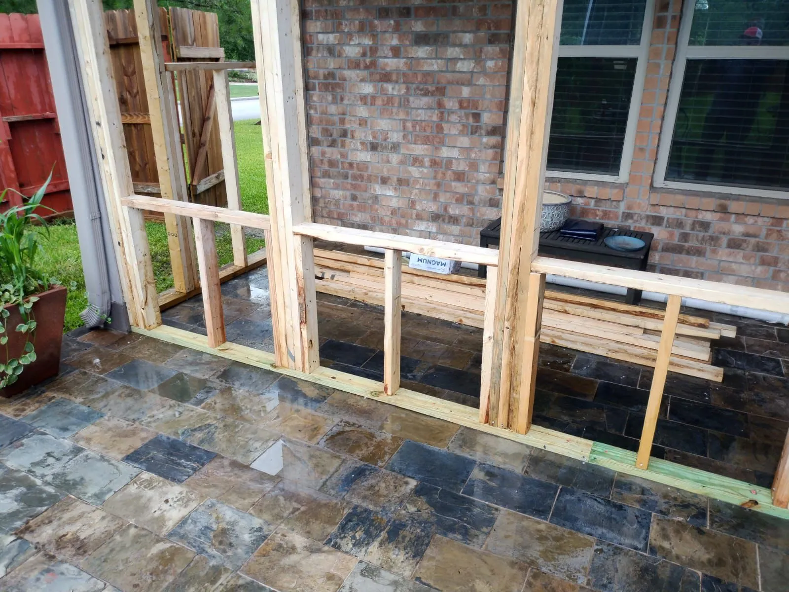Partially constructed wooden frame structure on a wet stone tiled patio next to a brick wall with stacked lumber and patio furniture in the background.