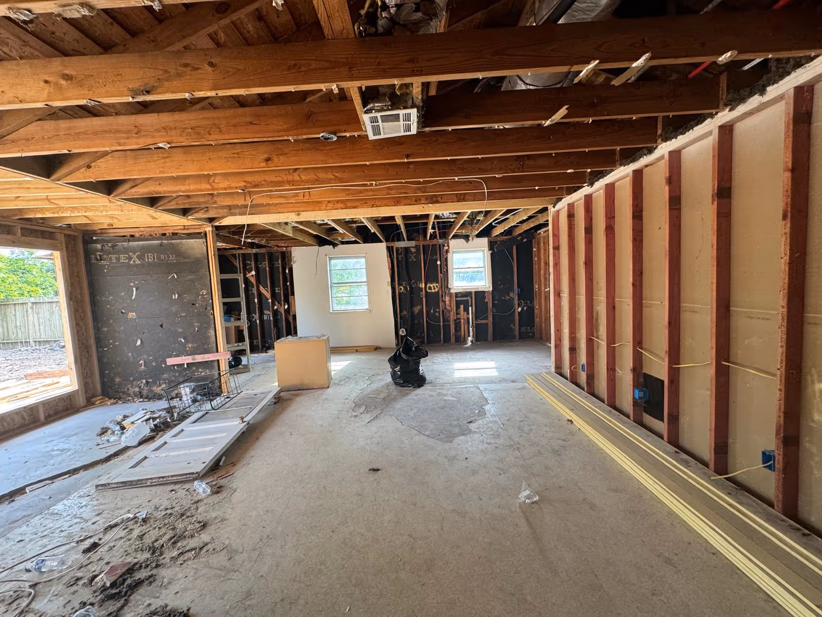 Interior of a house under renovation showing exposed wooden ceiling beams, partially framed walls, and a concrete floor with a trench cut for foundation work.
