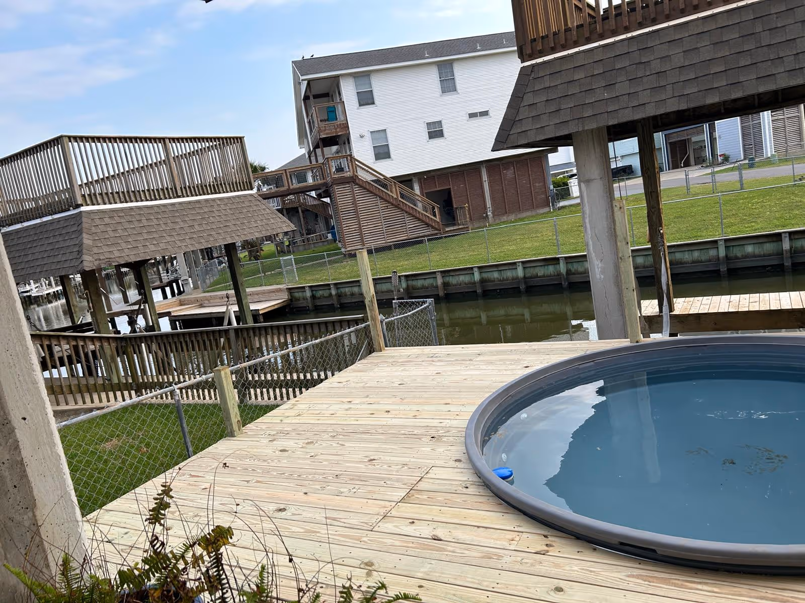 Wooden deck with a hot tub next to a canal, with houses and boat docks in the background.