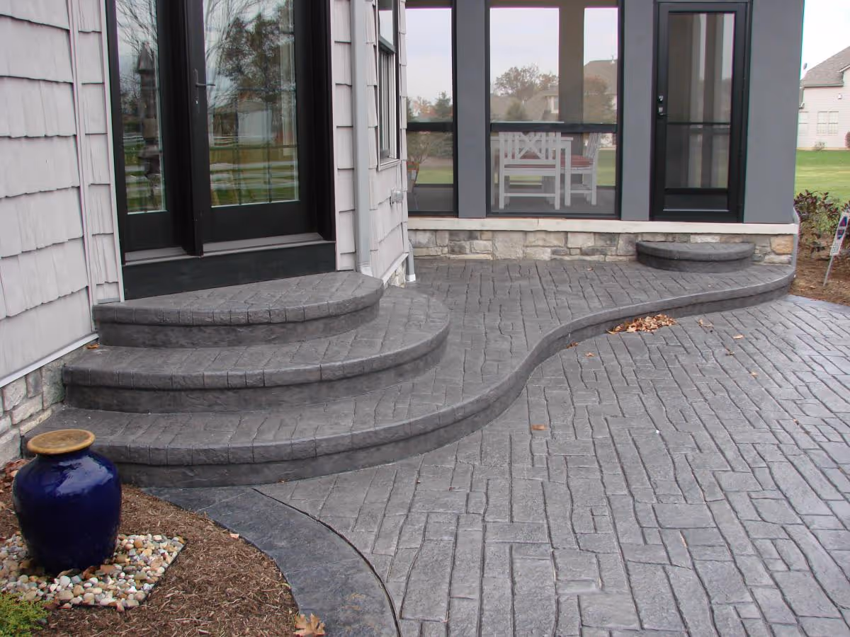 Curved gray stamped concrete patio with steps leading to black-framed glass doors and a blue ceramic planter on mulch.