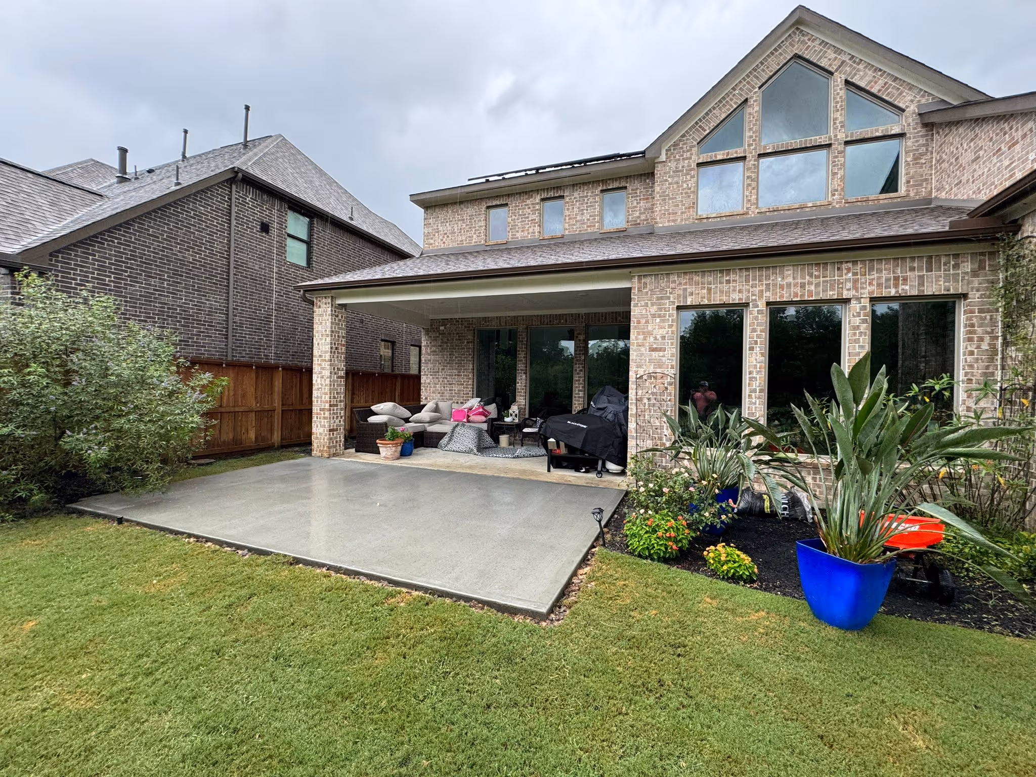 Partially constructed wooden frame structure on a wet stone tiled patio next to a brick wall with stacked lumber and patio furniture in the background.