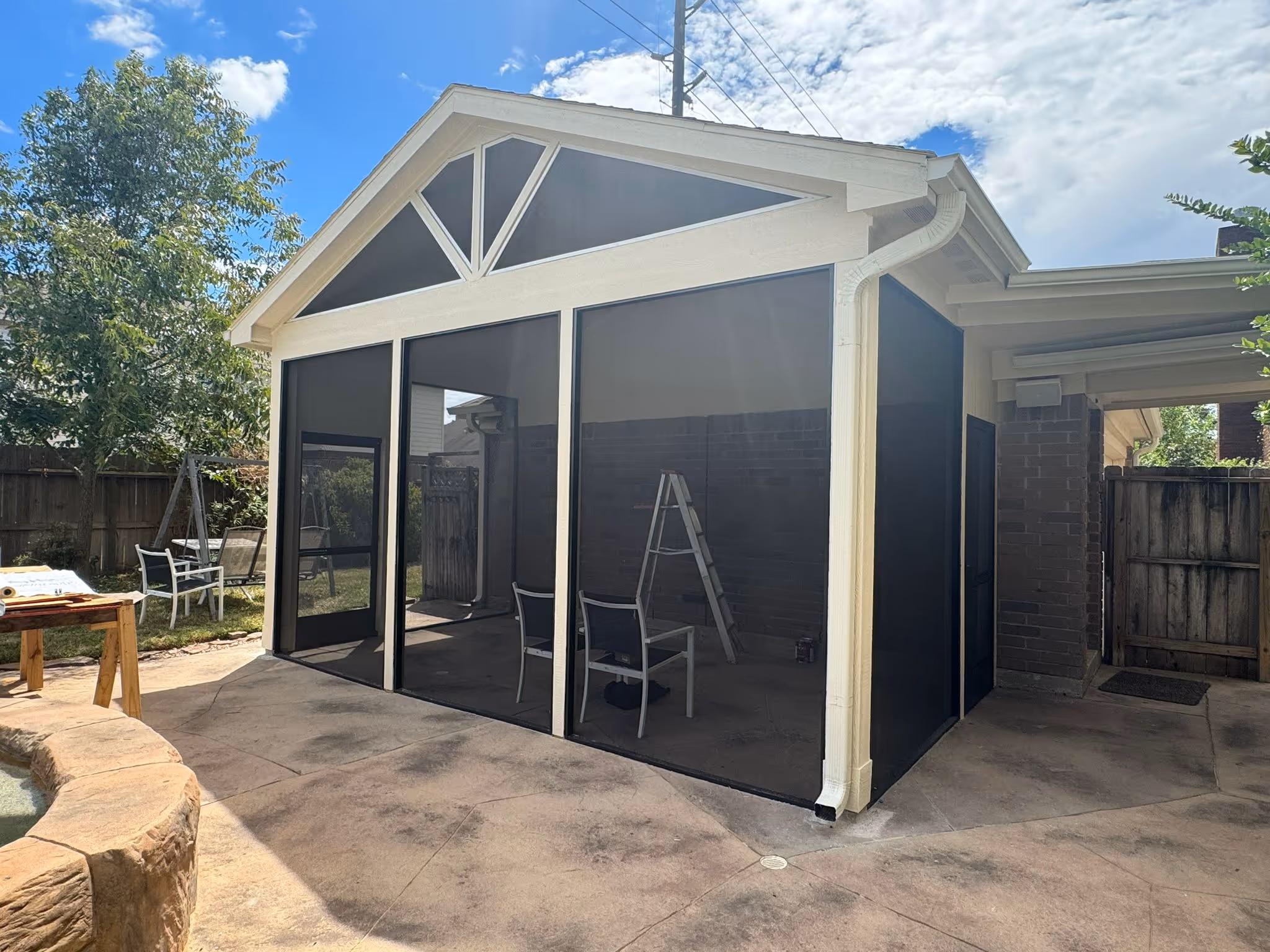 Screened patio enclosure with a pitched roof, two chairs, and a ladder inside, next to a stone-edged pool and backyard fence.