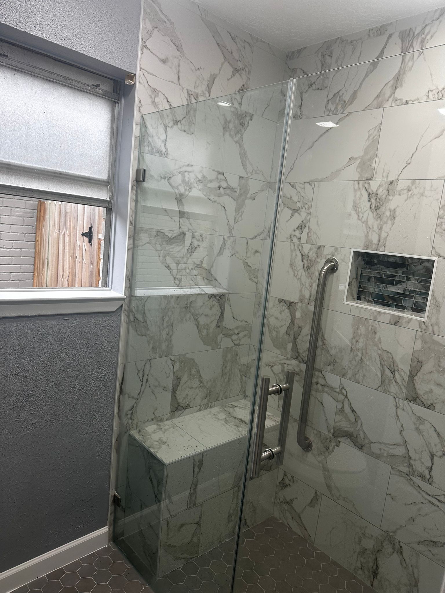 Walk-in shower with gray marble-look tile, a glass panel, a built-in bench, and a grab bar.