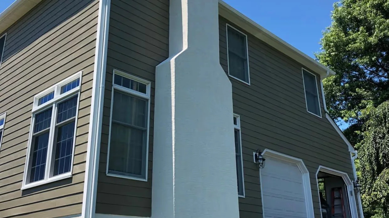 Two-story home with olive-green lap siding, white window trim and a white stucco chimney beside the garage.