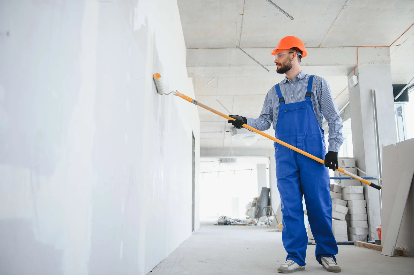 Painter in blue overalls and a hard hat rolling paint onto a white interior wall.
