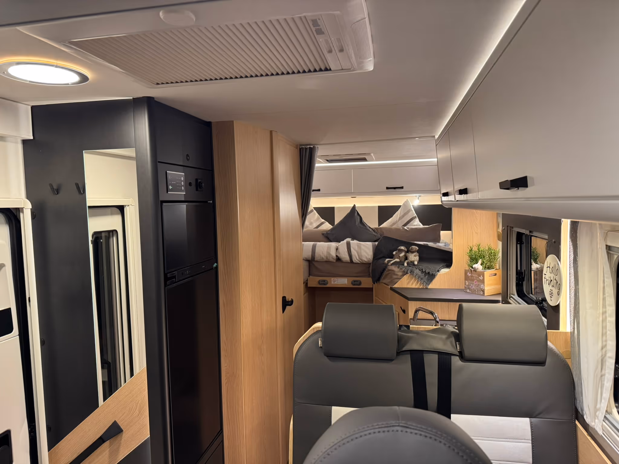 Innenraum eines modernen Wohnmobils mit grauen Sitzen, Küchenbereich, Bett mit Kissen und Dekoration.
