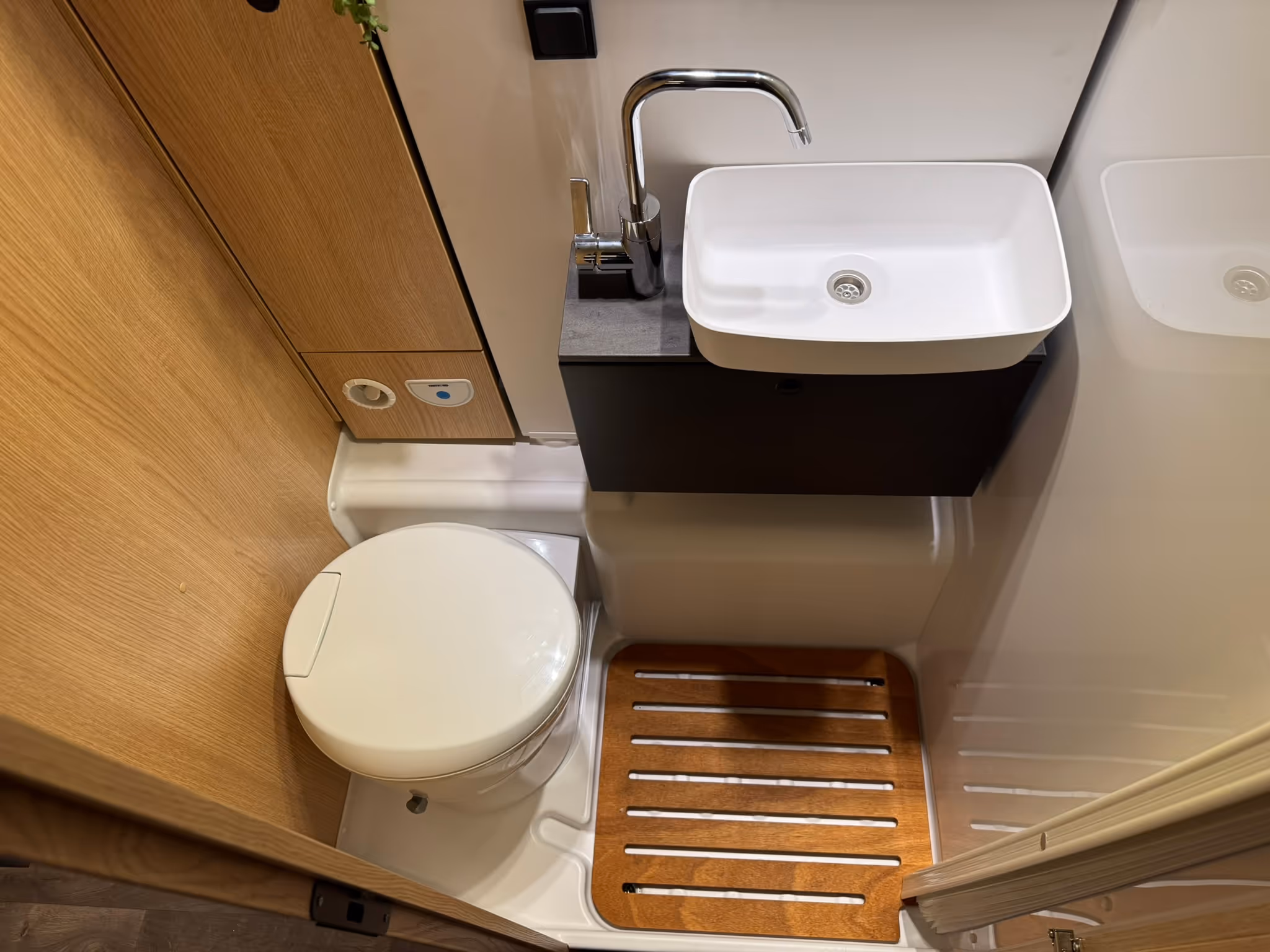 Kompaktes Wohnmobil-Badezimmer mit weißer Toilette, rechteckigem Waschbecken und Dusche mit Holzlattenboden.
