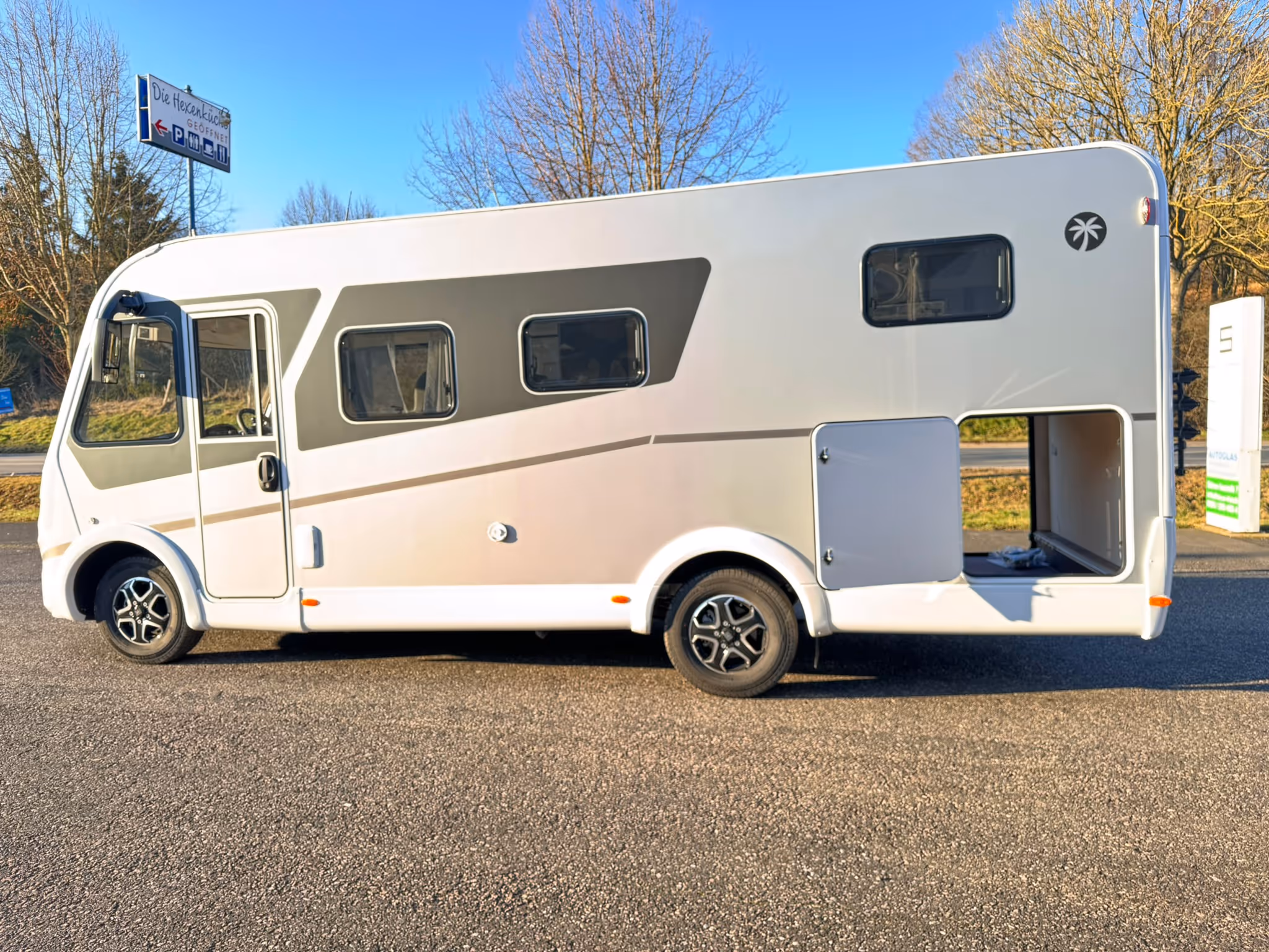 Weißes Wohnmobil mit aufgesetztem grauen Streifen und geöffnetem Heckstauraum, geparkt auf Asphalt unter klarem Himmel.