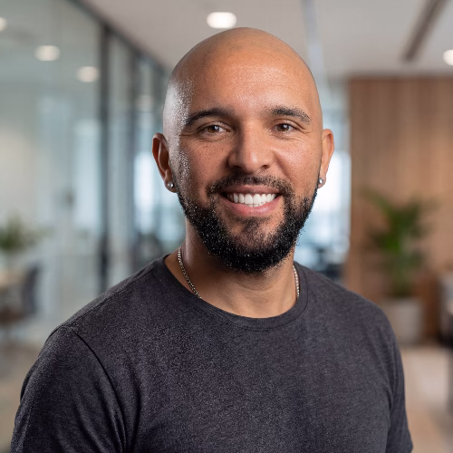 Smiling bald man with a beard wearing a dark gray shirt in a modern office setting.