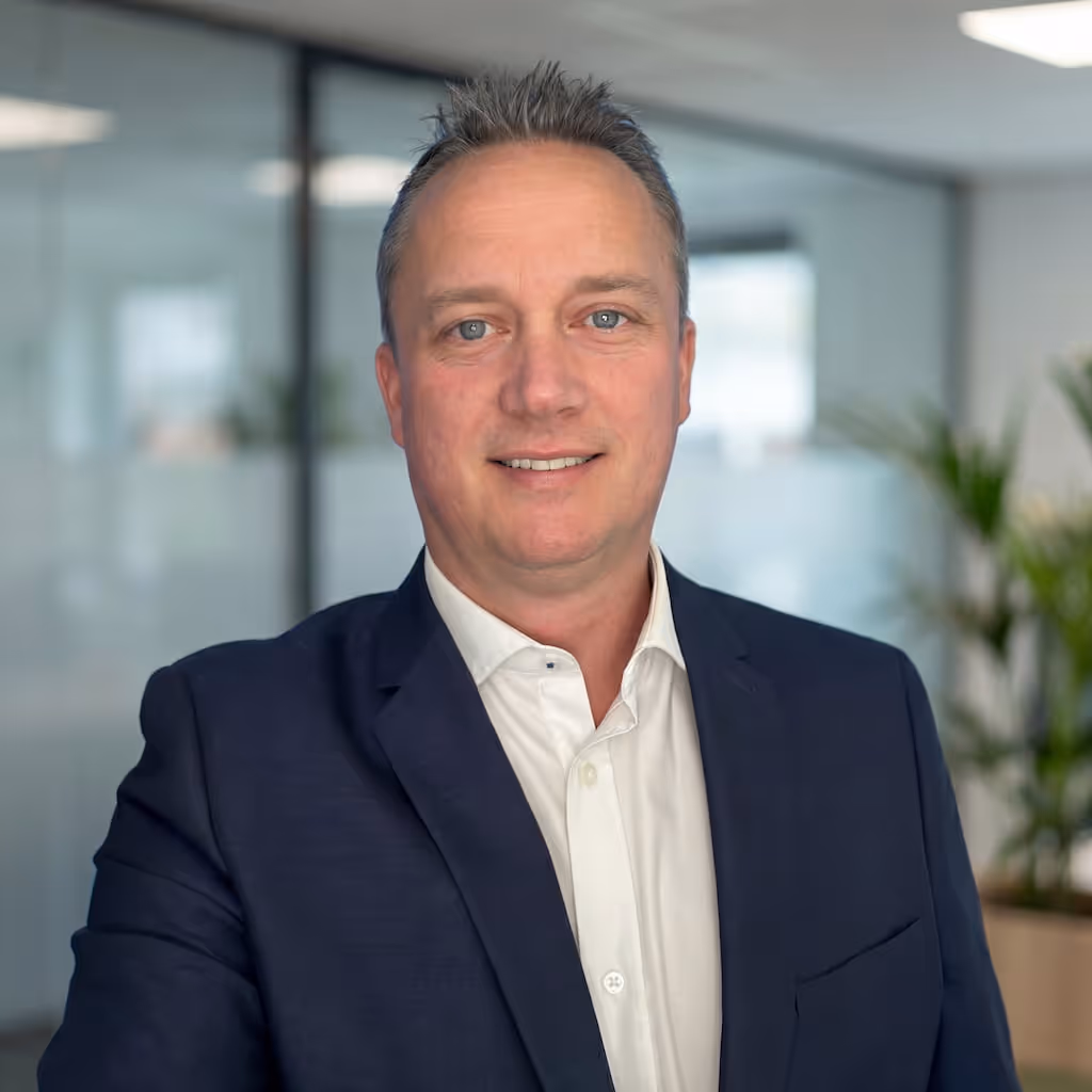 Man in a navy blue blazer and white shirt smiling in a modern office with glass walls and plants.