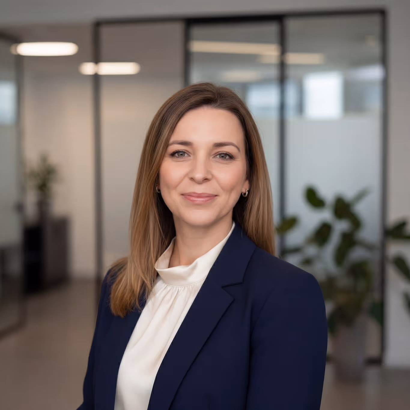 Smiling woman with light brown hair wearing a navy blazer and white blouse in a modern office setting.