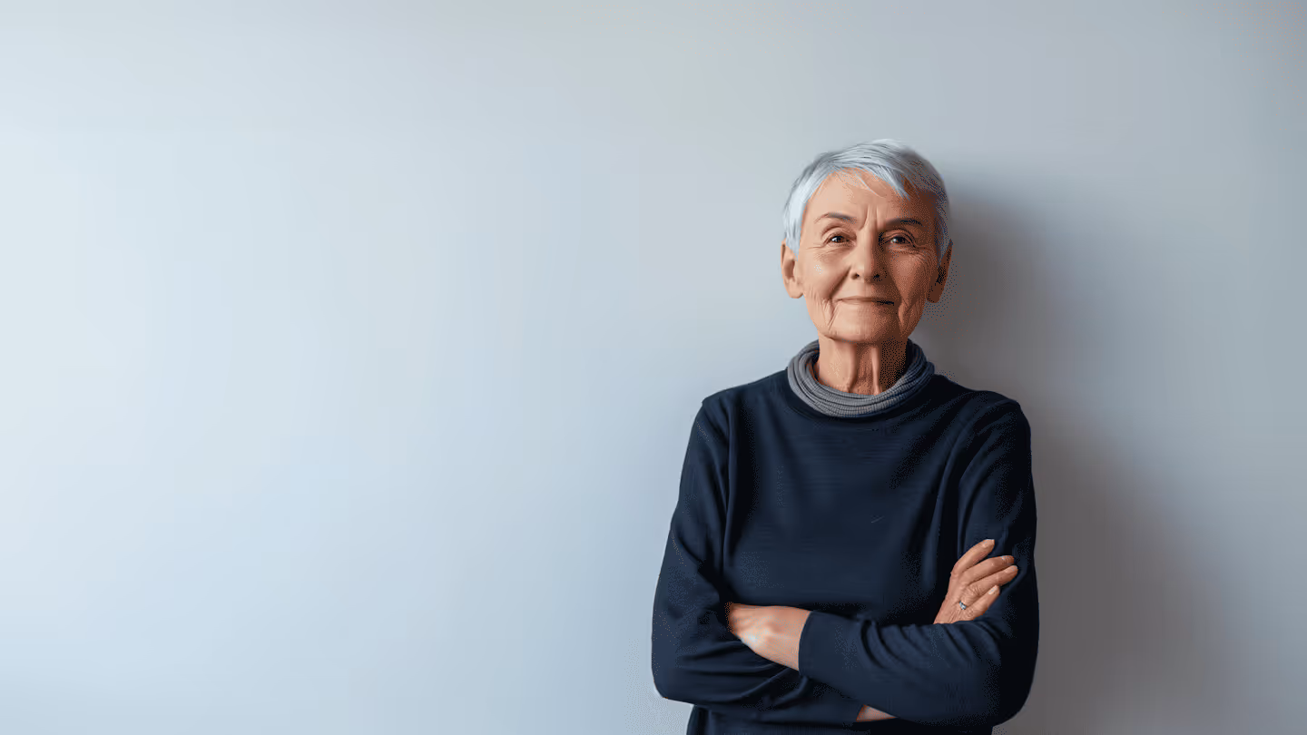 Ältere Frau mit kurzen grauen Haaren und verschränkten Armen vor hellgrauer Wand.