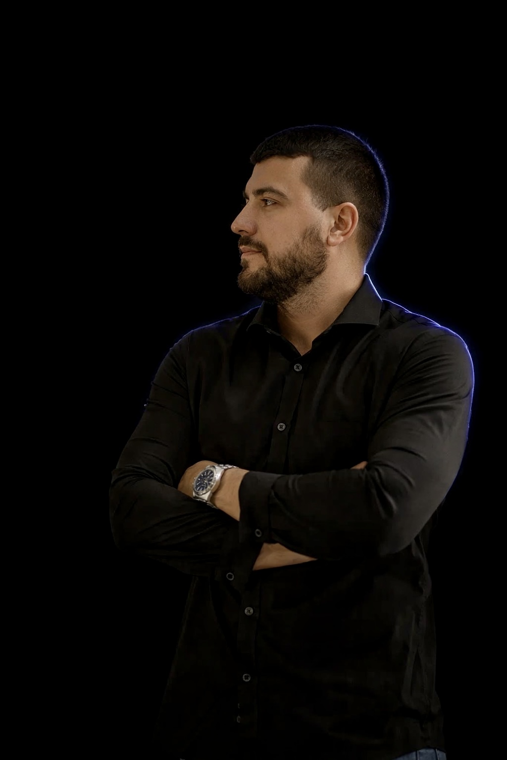 Bearded man wearing a black shirt and silver watch standing with arms crossed looking to the side against a black background.
