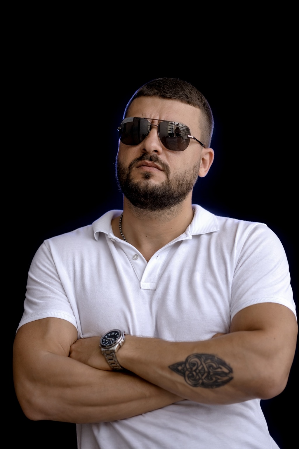 Bearded man wearing sunglasses and a white polo shirt with folded arms showing a tattoo on his forearm.