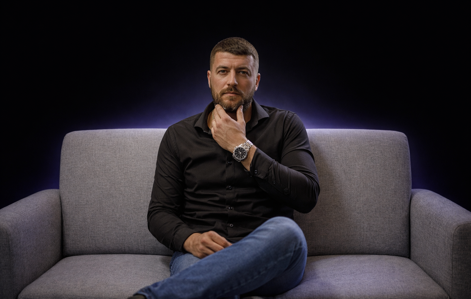 Man with short hair and beard wearing a black shirt and watch, sitting cross-legged on a gray sofa against a dark background with purple backlight.