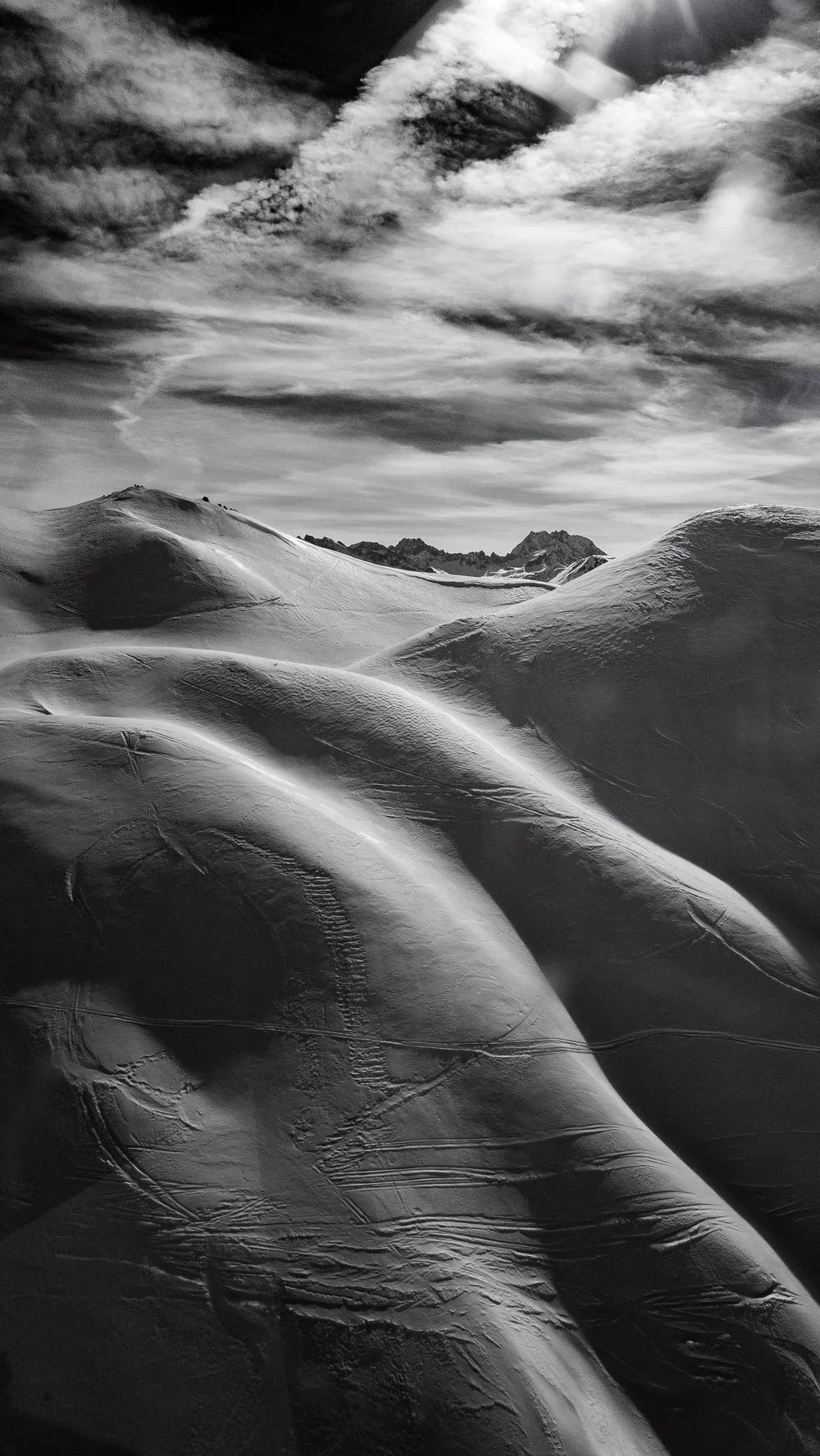 Black and white photo of snow-covered mountain slopes under a cloudy sky with ridges in the background.