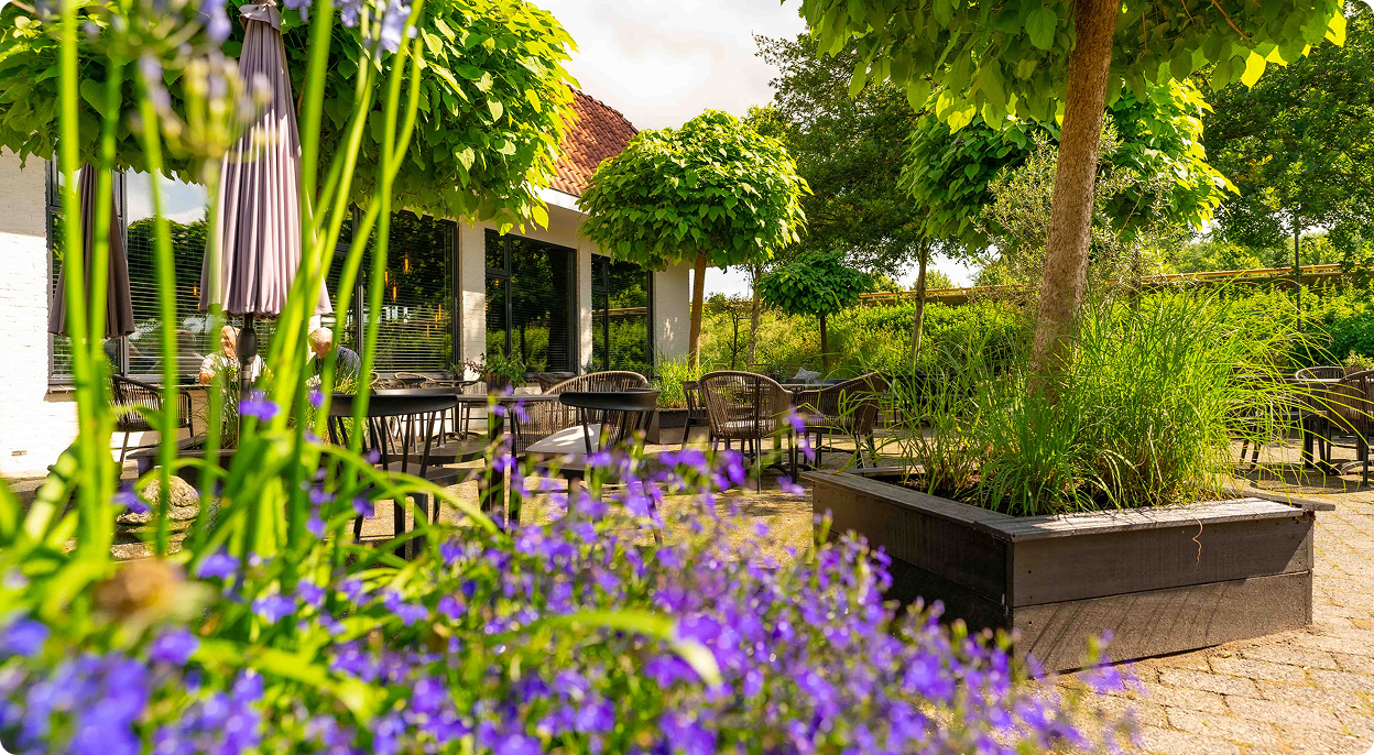 Terras met tafels en stoelen omgeven door groen en paarse bloemen in een zonnige tuin.