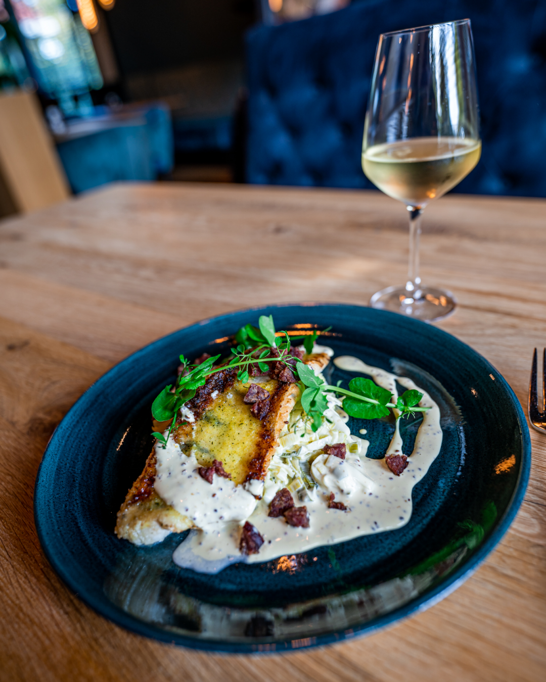 Een bord met krokant gebakken visfilet bedekt met saus, gegarneerd met kleine stukjes spek en groene kruiden, geserveerd met een glas witte wijn op een houten tafel.