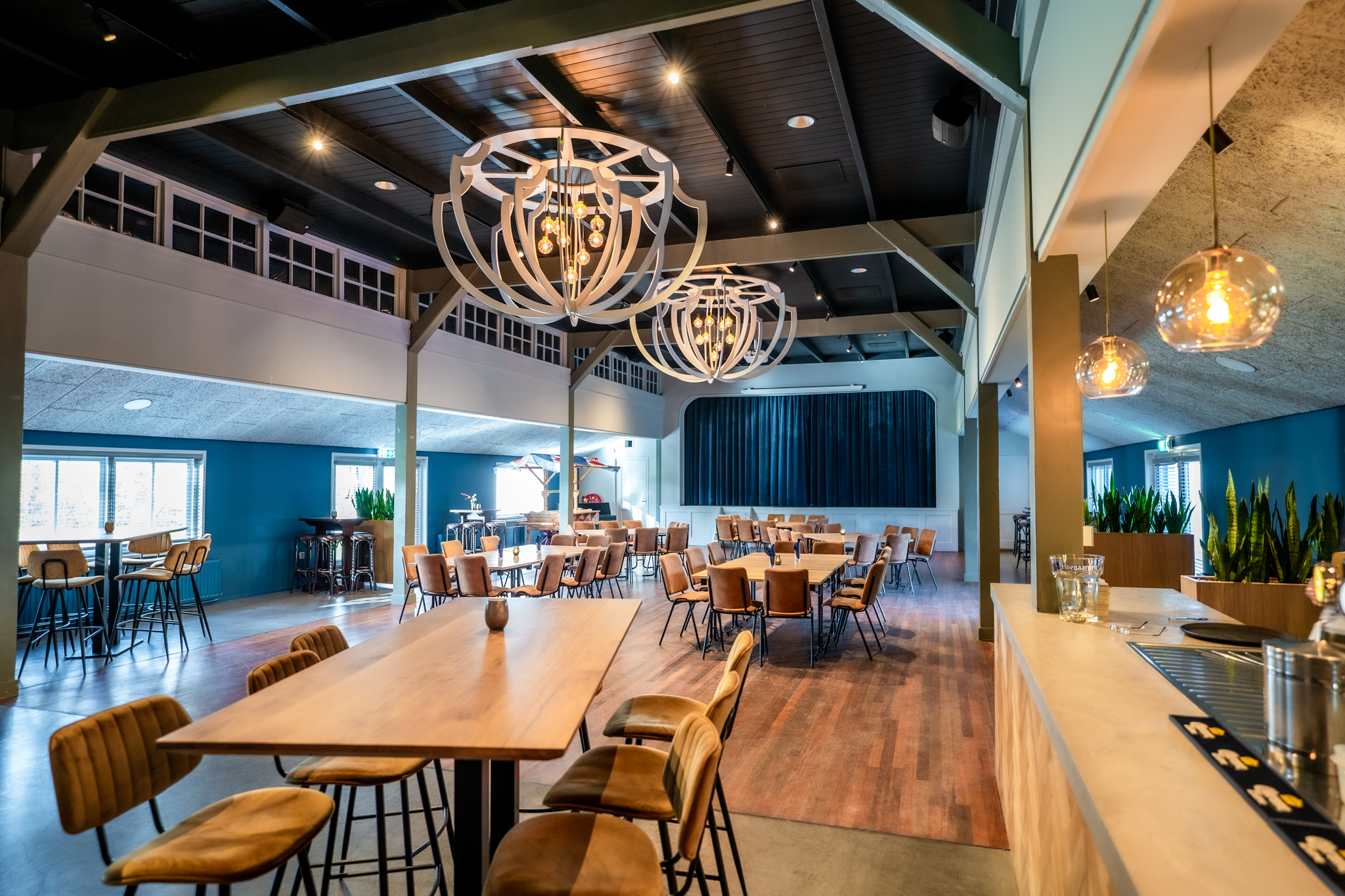 Modern restaurantinterieur met houten tafels, bruine stoelen, hanglampen en een podium met een donkerblauwe gordijn.