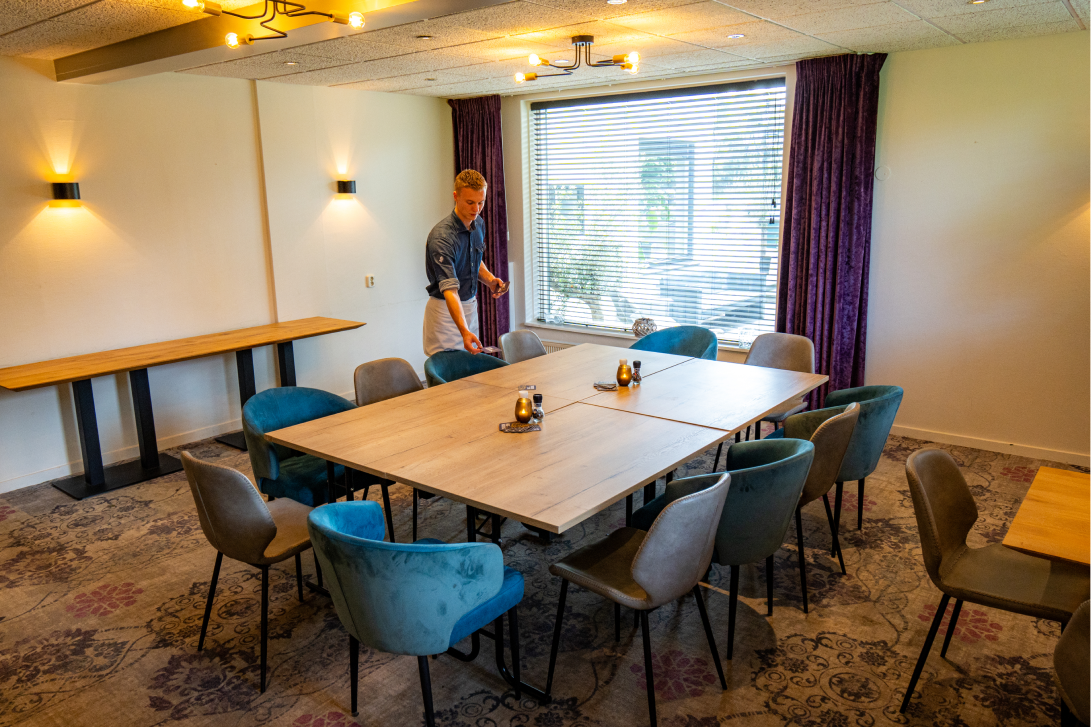 Een man die een stoel rond een grote vierkante vergadertafel in een sfeervolle kamer met blauw en grijs gestoffeerde stoelen en een groot raam met jaloezieën, klaarzet.