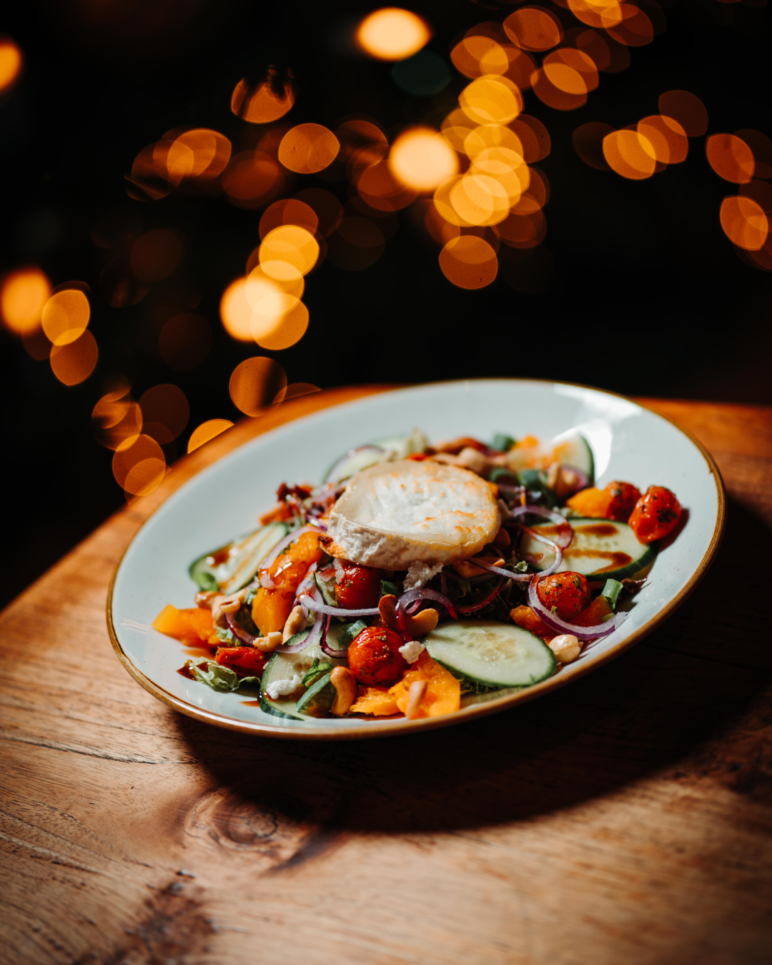 Salade met gegrilde geitenkaas, komkommer, cherrytomaatjes, rode ui en noten op een witte schaal op een houten tafel met zachte, warme verlichting op de achtergrond.