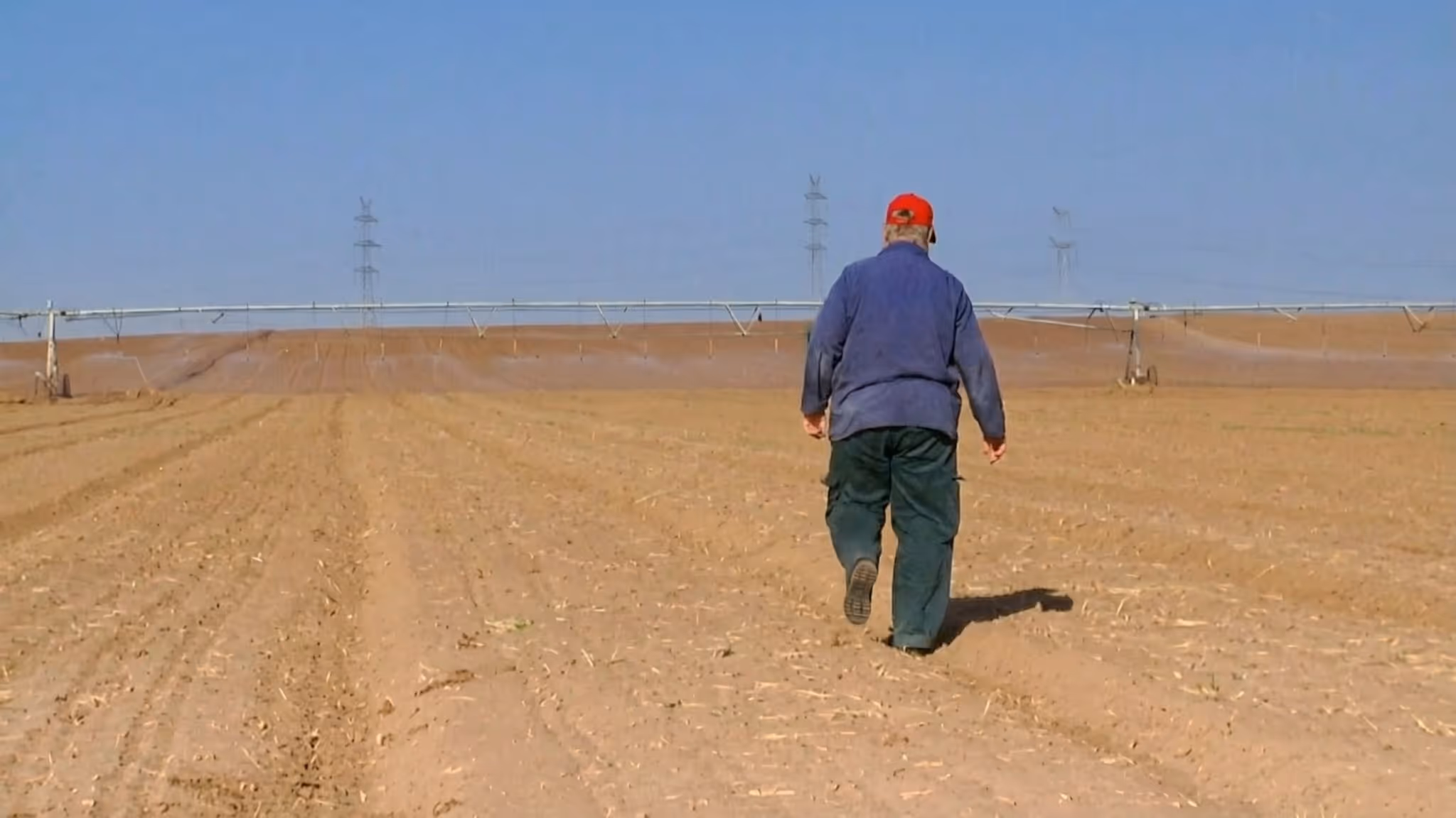 An elderly man walking alone across a vast plowed agricultural field with irrigation equipment, from the documentary series Sixty