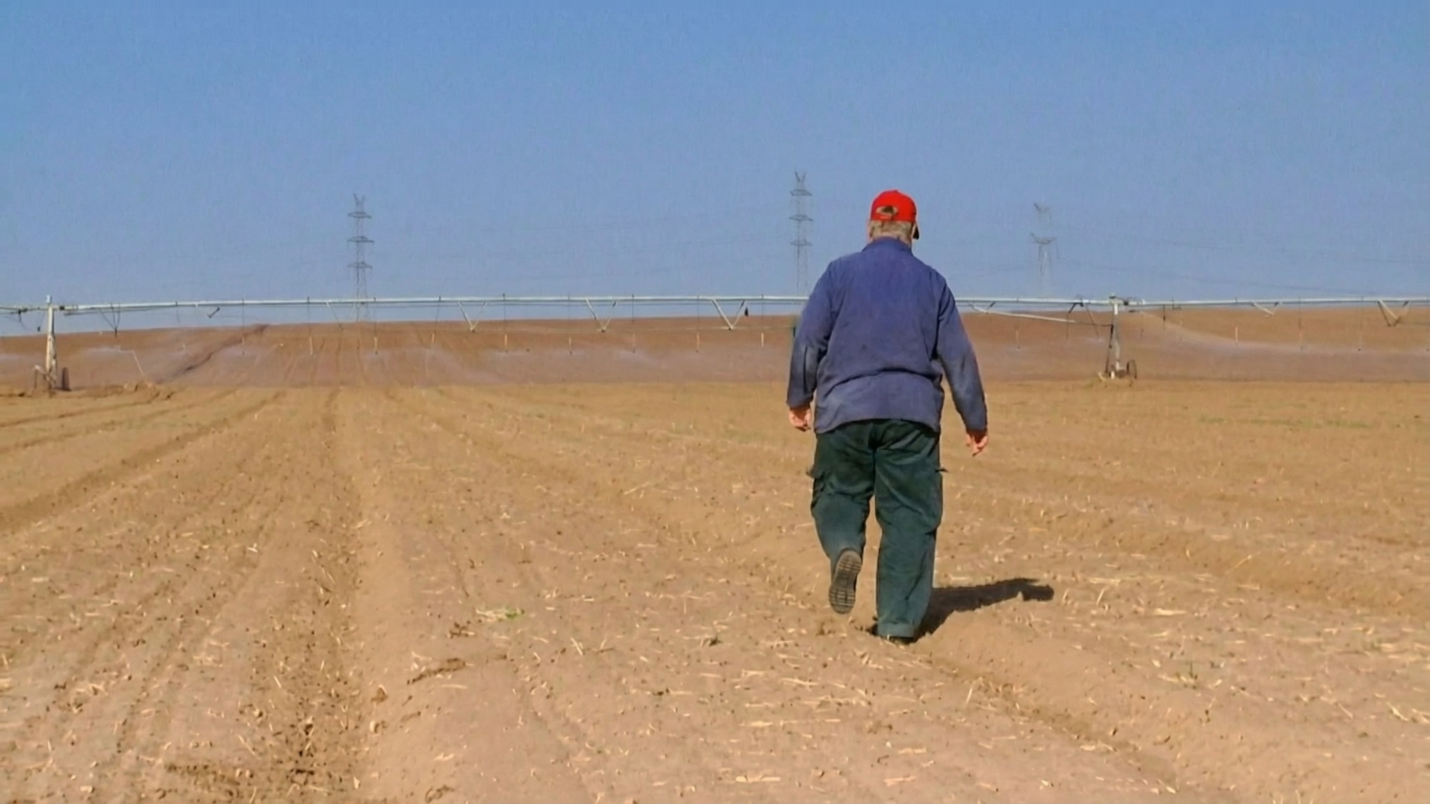 An elderly man walking alone across a vast plowed agricultural field with irrigation equipment, from the documentary series Sixty