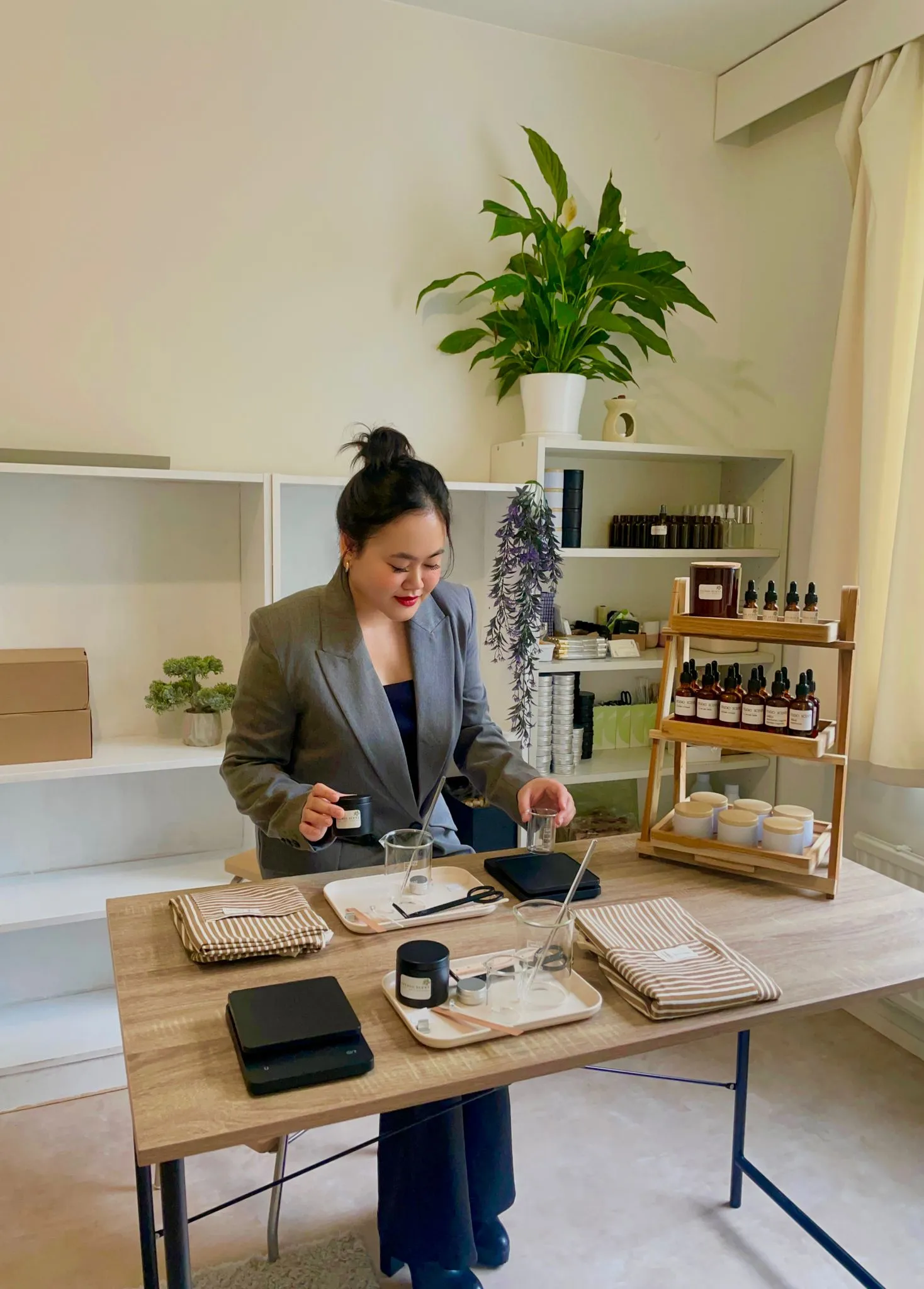 Woman in gray blazer sitting at a wooden table with cosmetic jars and tools in a light room with shelves and a potted plant.