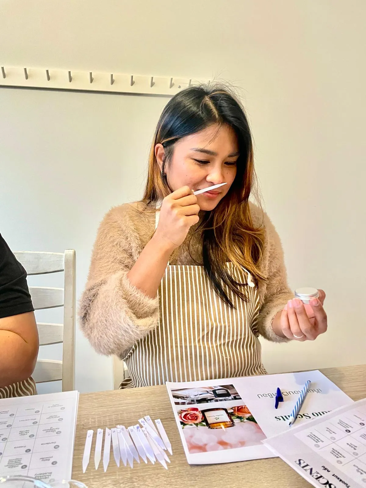 Woman in striped apron smelling a fragrance strip while holding a small container, sitting at a table with scent testing strips and papers.