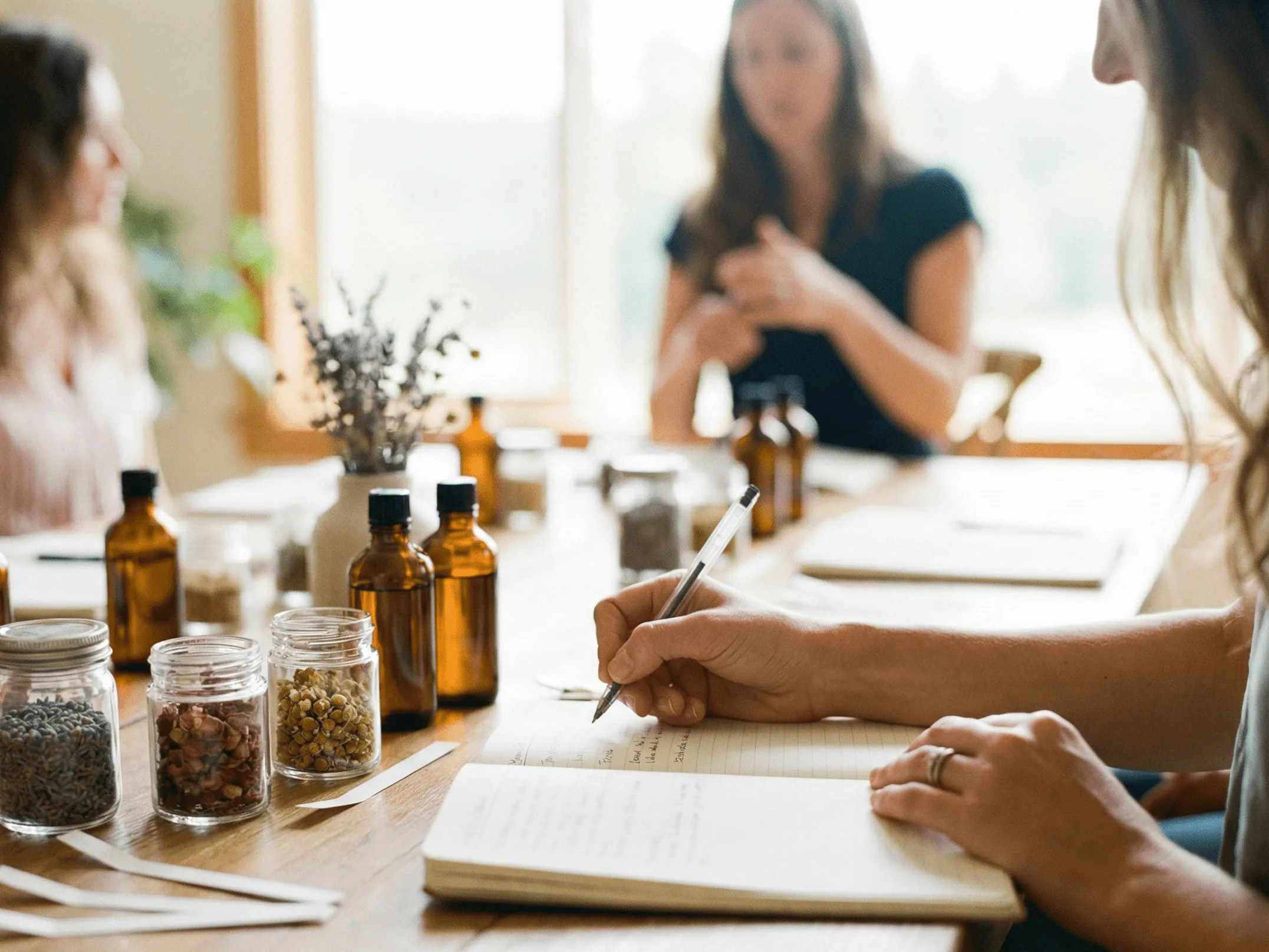 Person writing notes in a notebook at a wooden table with amber bottles, jars of dried herbs, and two blurred people in the background.