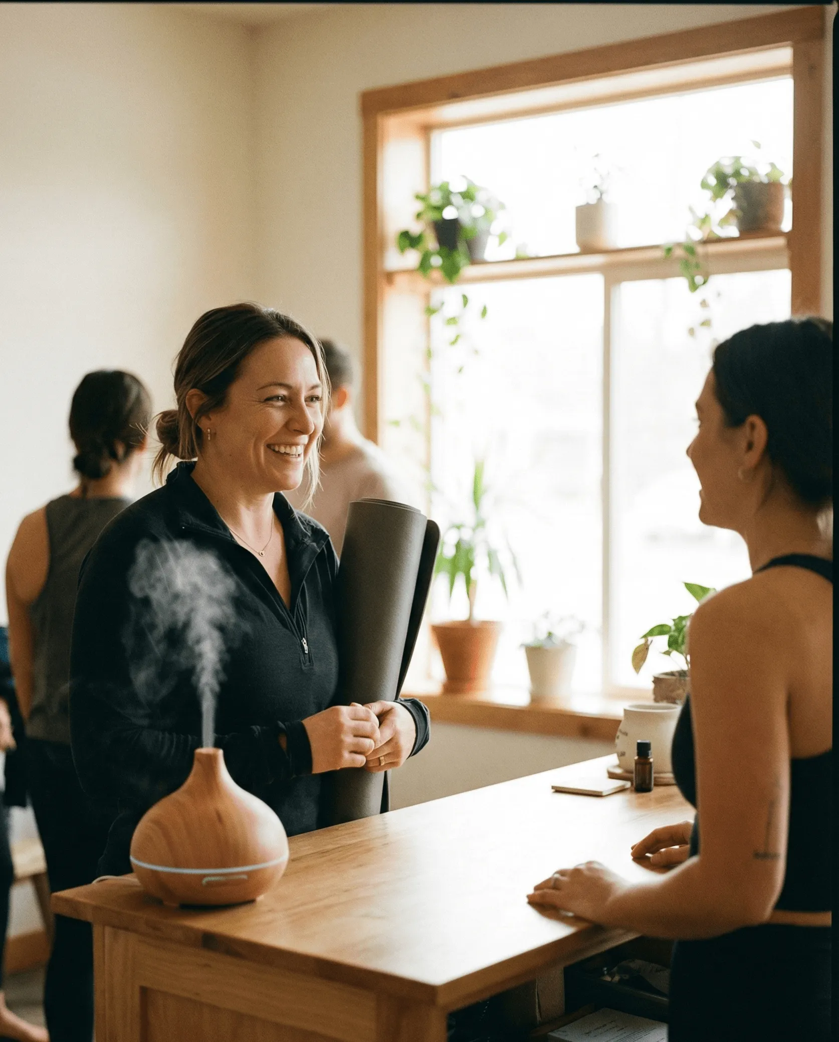 Smiling woman holding a yoga mat talks to another woman across a wooden counter with a diffuser emitting vapor nearby.