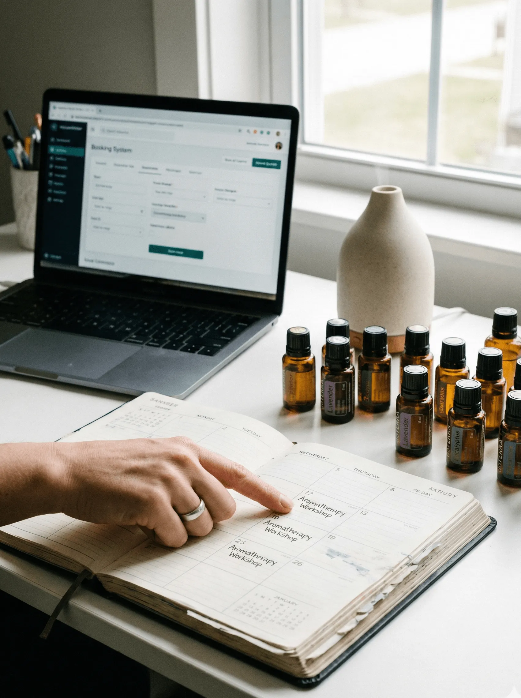 Person pointing at an entry labeled Aromatherapy Workshop in a planner with essential oil bottles and a laptop on a white desk near a window.