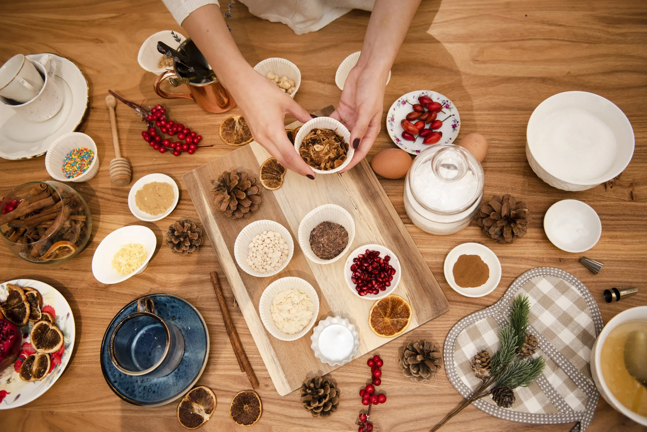Top view of a wooden table with baking ingredients including eggs, sugar, spices, pomegranate seeds, dried orange slices, pine cones, and two hands holding a cup with granola.